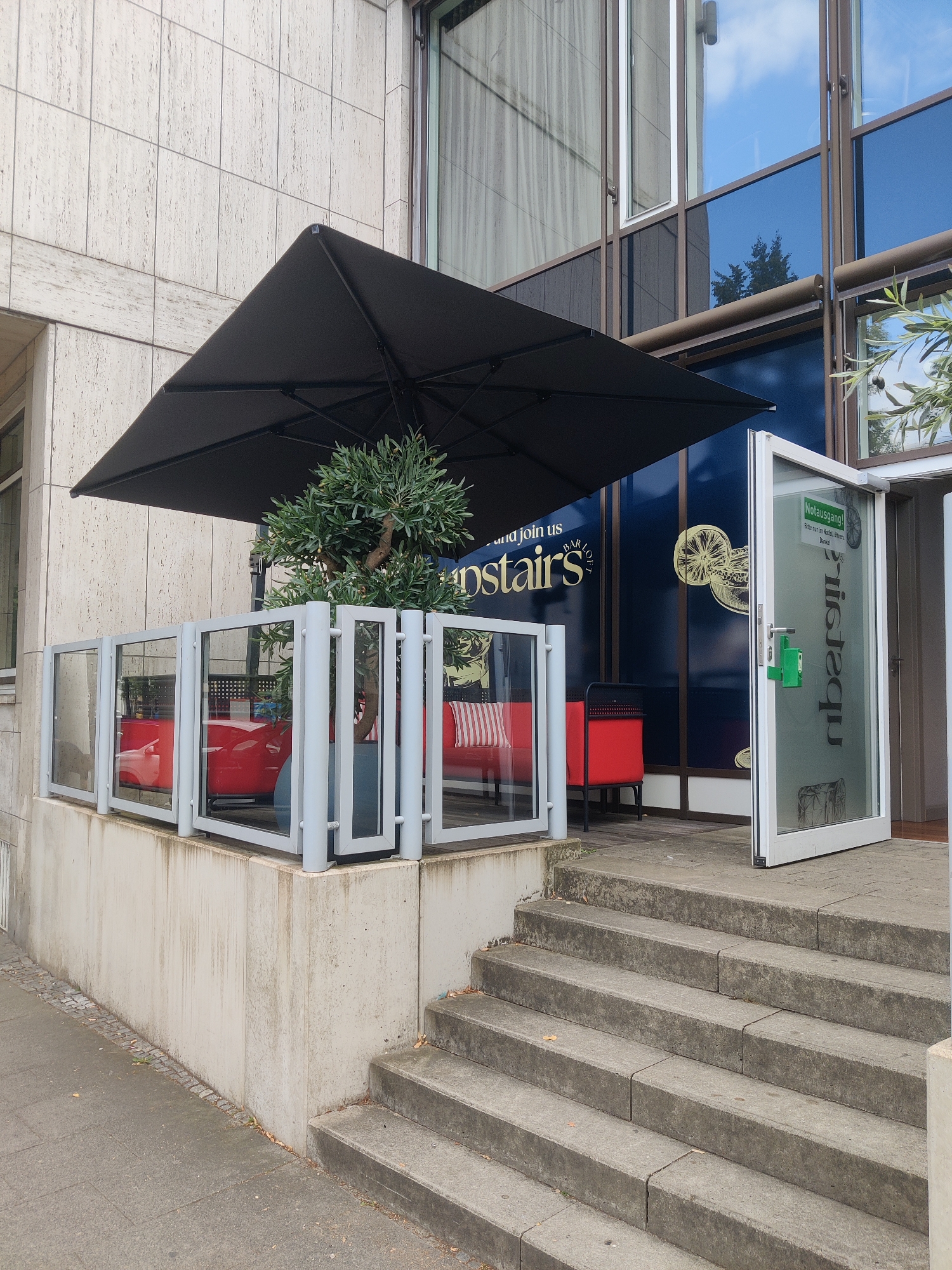 This image captures an outdoor seating area of a establishment, likely a bar or restaurant, on a cloudy day. The scene is set in front of a modern building with a facade of light-colored stone tiles and dark blue tinted windows.
Dominating the foreground is a large, open black umbrella, providing shade over a section of the patio. Below the umbrella, a contemporary seating area is visible, enclosed by a glass and metal railing. Inside this enclosure, two red benches with white striped cushions are placed, hinting at a place for patrons to relax. A small, potted olive tree stands in the center of the seating area, adding a touch of greenery.
To the right of the patio, a glass door stands ajar, leading into the building. A sign on the door reads "Notausgang!" (Emergency Exit!) in bold, red letters, with smaller text below indicating "Bitte nur in Notfall öffnen" (Please open only in case of emergency) and "Danke!" (Thank you!). The frosted glass of the door features a vertical text inscription that appears to be backwards, reading "aquaribs", possibly a business name or part of a logo.
The foreground also shows a concrete staircase leading down to a paved sidewalk. The overall impression is a modern, urban setting, likely in Germany, with a functional and inviting outdoor space. The lighting suggests it is daytime, but the overcast sky hints at a cooler or possibly rainy atmosphere. No people are visible in the image.