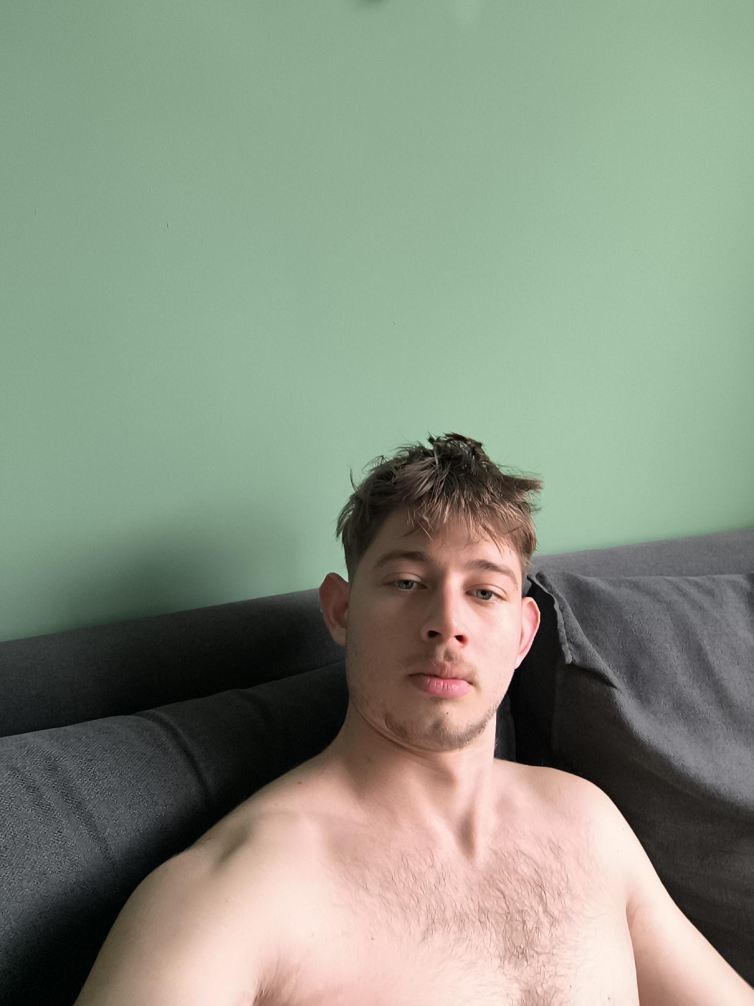 A shirtless man is lying on a gray couch against a mint green wall. The man has light skin, brown hair styled messily, and blue eyes. He is looking directly at the camera. The environment suggests an interior setting. There is no verifiable information to indicate the location in Sofia, Bulgaria.