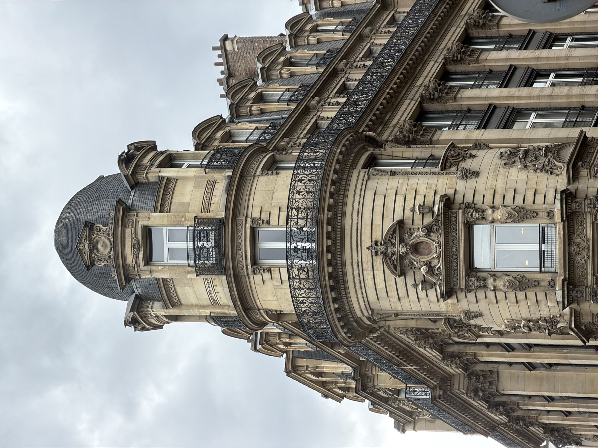 A low-angle shot showcases the architecture of a Parisian building. The building is intricately designed with a dome on the corner, multiple windows, and decorative iron balconies. The sky is overcast.