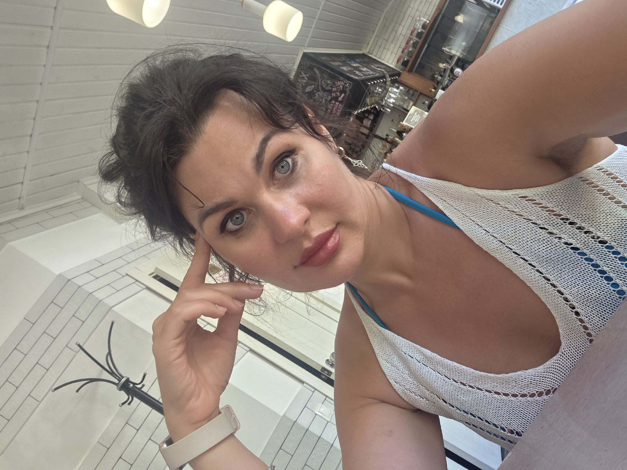 A woman with dark hair pulled back, blue eyes, and pink lipstick takes a selfie in a cafe in Sochi, Russia. She is wearing a white crochet top over a blue bra, and a white smartwatch on her left wrist. Her right hand is raised to her temple. The background shows a white tiled wall, a coat rack, and a ceiling with exposed beams and lighting fixtures. A shelf with bottles and jars is visible in the distance.