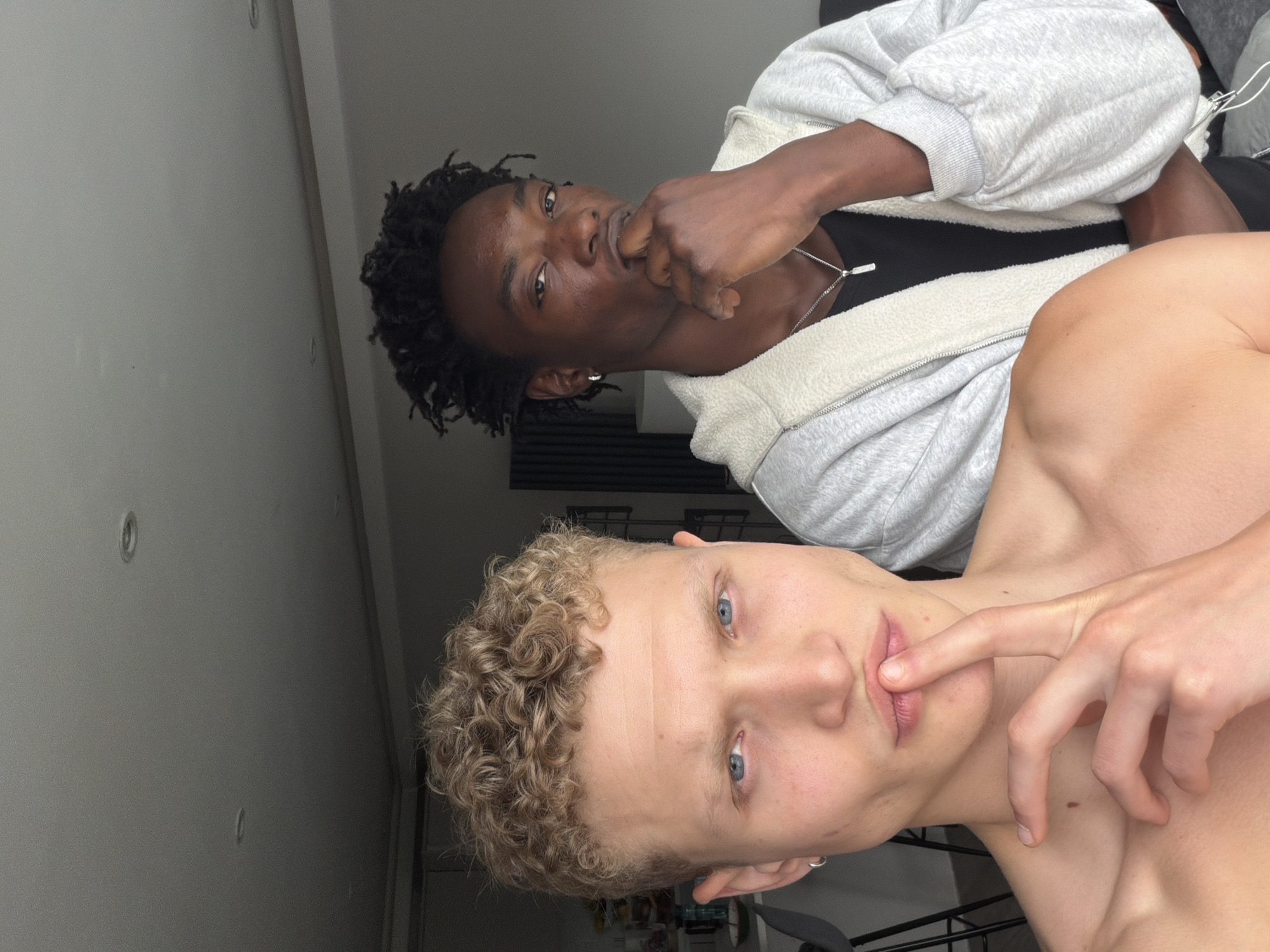 The image displays two young adult males posing in an indoor residential setting, likely an apartment in London, United Kingdom.
In the foreground, a light-skinned male with short, tightly curled blonde hair and blue eyes looks directly at the viewer. He is bare-chested, exposing his shoulders and upper torso. His right index finger is raised to his lips in a "shhh" gesture. A silver hoop earring is visible in his left ear.
Behind and to the right of the first individual stands a dark-skinned male with short, dark dreadlocks/twists and dark eyes, also looking directly at the viewer. He wears a black t-shirt beneath an open, light grey, fleece-lined hooded jacket. His left hand is raised to his chin, with his index finger and thumb gently gripping his lower face. A silver chain necklace with a small, rectangular silver pendant hangs around his neck, and a silver hoop earring is visible in his left ear.
The background reveals a modern, minimalist interior. To the left, part of a kitchen or dining area is visible with light-colored countertops, some glassware, and the back of a dark bar stool. The walls are light grey, and the ceiling features two recessed circular lights. In the mid-background, a dark, vertical panel radiator is mounted on the wall. The overall lighting is soft and even, suggesting indirect light sources.
