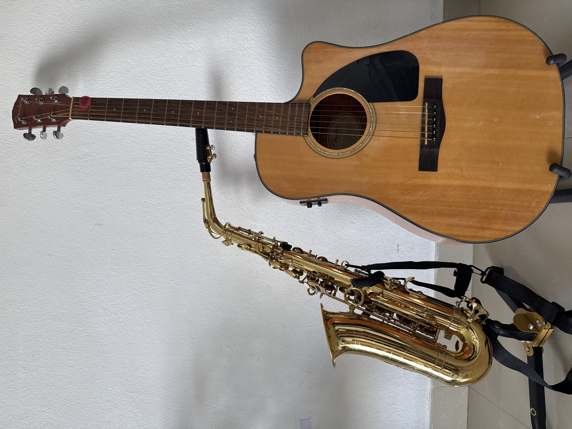 The image shows a guitar and a saxophone on a stand against a white wall. The guitar is an acoustic-electric model with a light-colored wood finish and a black pickguard. A red guitar pick is attached to the headstock. The saxophone is brass-colored and has a black neck strap attached. The stand is black and holds both instruments. The background is a plain white wall. The location is identified as Baan Chaweng Noi, Thailand, but no elements in the image independently verify this location. No people are visible.
