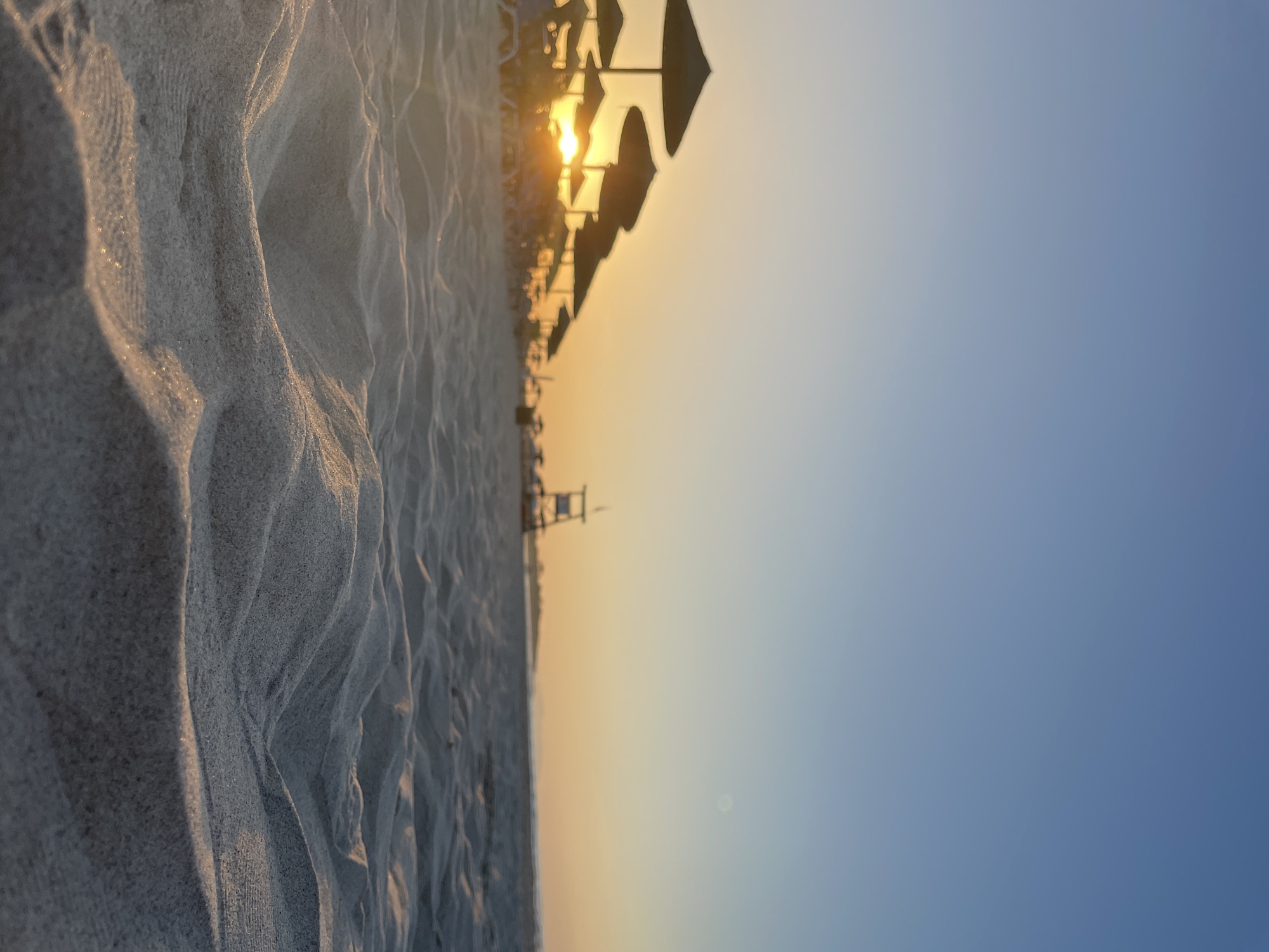 An extreme close-up shot of rippled sand dunes on a beach in Greece. The sun is setting, casting a warm glow over the scene. In the distance, a row of beach umbrellas and lounge chairs are visible, as well as a lifeguard tower. The sky is a clear blue, with a gradient of orange and yellow as it meets the horizon. The sand is fine and light-colored, with small shadows and highlights creating a textured appearance. The overall mood is peaceful and serene.