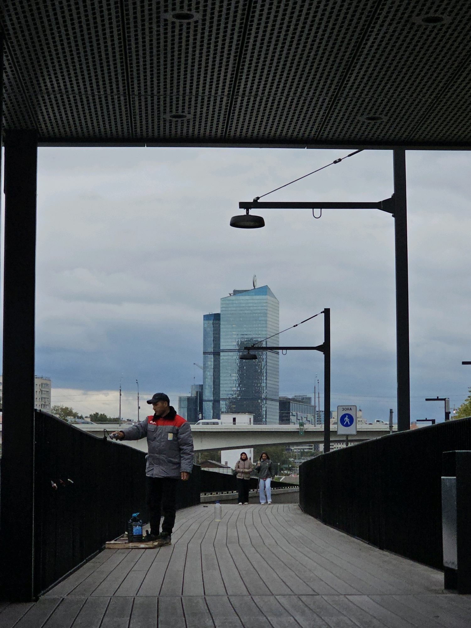 The image depicts an outdoor scene in Moscow, Russia, under a cloudy sky. In the foreground, a man wearing a gray and red jacket and a black cap is standing on a wooden walkway, holding a brush and appearing to be painting a black railing. Beside him, on a wooden pallet, are containers, one of which is blue.
Further down the walkway, two women are walking away from the camera. The woman on the left wears a light brown jacket and the woman on the right wears a dark jacket and light pants.
In the background, a modern, glass-facade skyscraper dominates the skyline. There is also a multi-lane highway overpass and other buildings. A sign with a pedestrian symbol and the Russian word "ЗОНА" (ZONA - Zone) is visible, indicating a designated pedestrian area. Streetlights and architectural elements of the walkway's canopy are also present. The overall atmosphere is urban and slightly overcast.