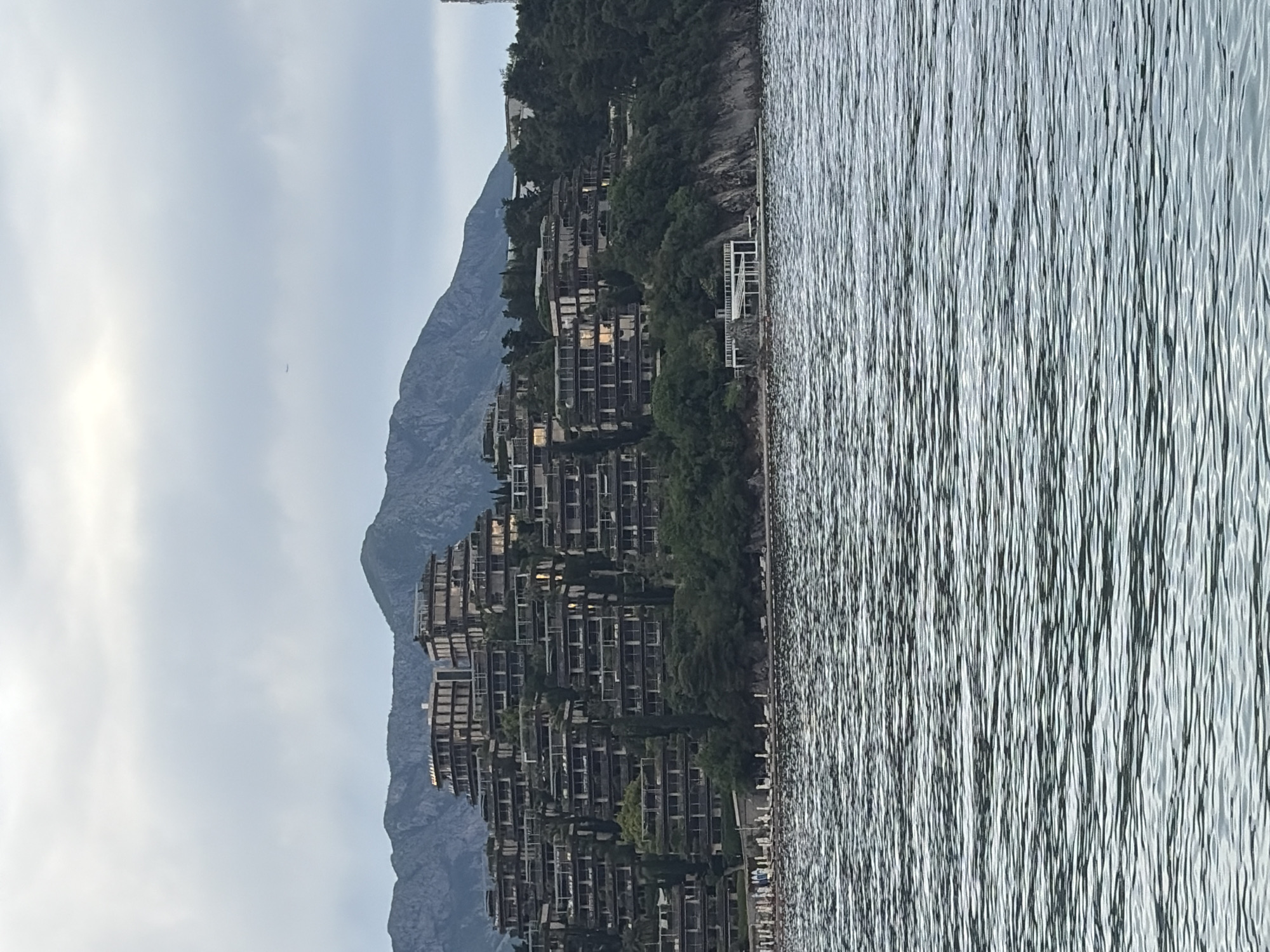 The image shows the Dukley Gardens residential complex located on the Zavala Peninsula in Budva, Montenegro. The buildings are constructed on a hillside, cascading towards the Adriatic Sea. The sea is visible on the right side of the image, with a rippled surface. Mountains rise in the background, partially obscured by the complex. The sky is overcast, with hints of sunlight. The buildings feature multiple tiers of balconies and are surrounded by trees and greenery. A white structure is situated near the water's edge.