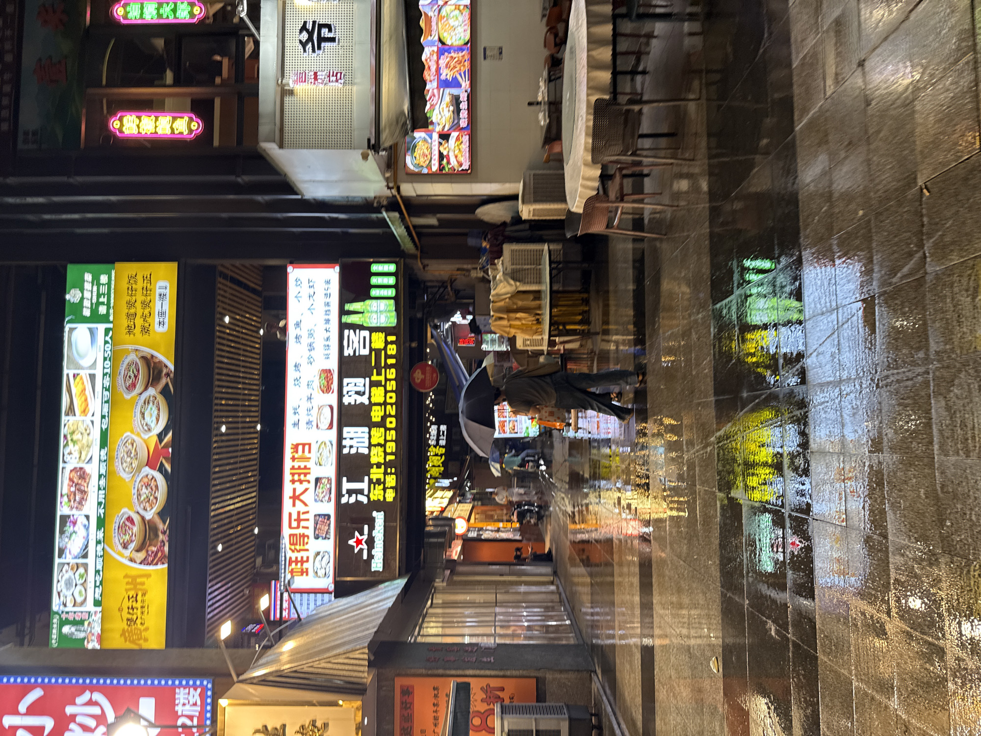 This image depicts a street scene at night in Haizhu District, China, with reflections of illuminated signs on the wet pavement. The street appears to be a commercial area with numerous restaurants and shops. A person is visible walking in the center of the frame, holding a black umbrella, suggesting it is raining or has recently rained. The reflections on the ground indicate a glossy, wet surface, likely due to rain. Various neon signs with Chinese characters are displayed, advertising food and businesses. Tables and chairs are set up outside some establishments, suggesting an outdoor dining area. The overall atmosphere is one of a bustling urban environment illuminated by artificial lights, with the wet conditions adding a distinct visual element.