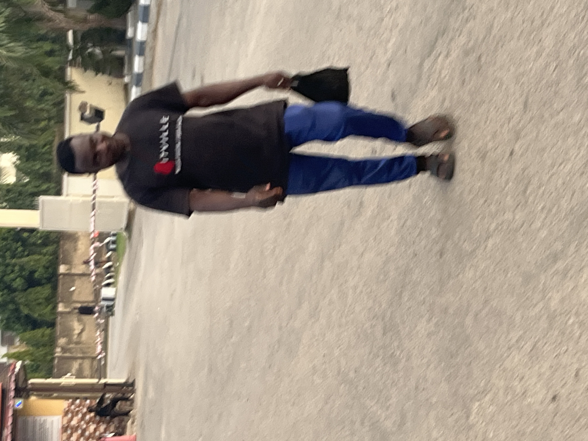A man is standing on a paved surface outdoors. He is wearing a black T-shirt with white writing and a red heart, blue pants, and sandals. He is holding a black hat in his right hand. In the background, there are trees and buildings, including a light-colored structure with a black and white barrier. Cars are parked along the side of the road. Based on the surroundings, the location is possibly in Abuja, Nigeria.