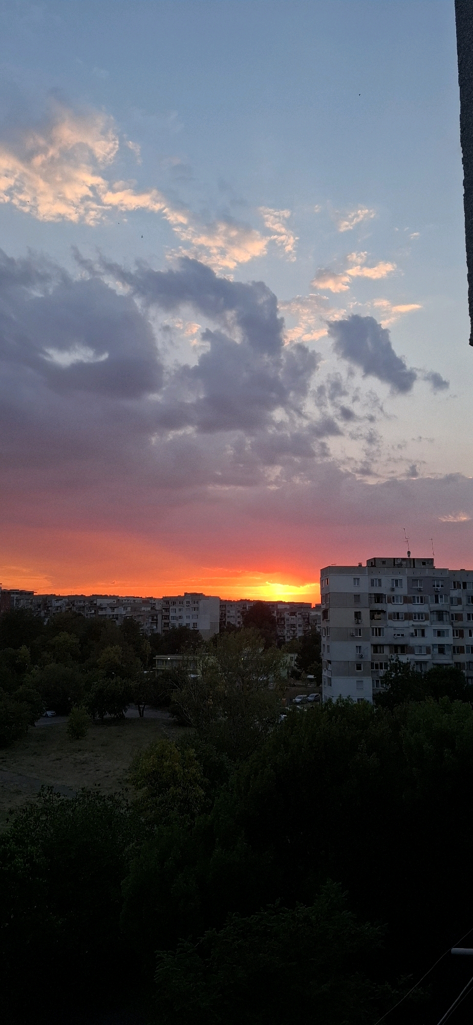 The image depicts a vivid sunset over a residential area in Sofia, Bulgaria. The sky is ablaze with hues of orange and pink, with clouds casting dramatic shadows. In the foreground, silhouetted trees and greenery create a dark contrast to the vibrant sky. Beyond the trees, apartment buildings, characteristic of Sofia's urban landscape, are visible. The architecture suggests a Soviet-era housing block, with multiple stories and balconies. A portion of a building, possibly a rooftop or balcony railing, juts into the frame from the right side, adding a sense of perspective. The overall scene captures a moment of natural beauty in an urban setting.