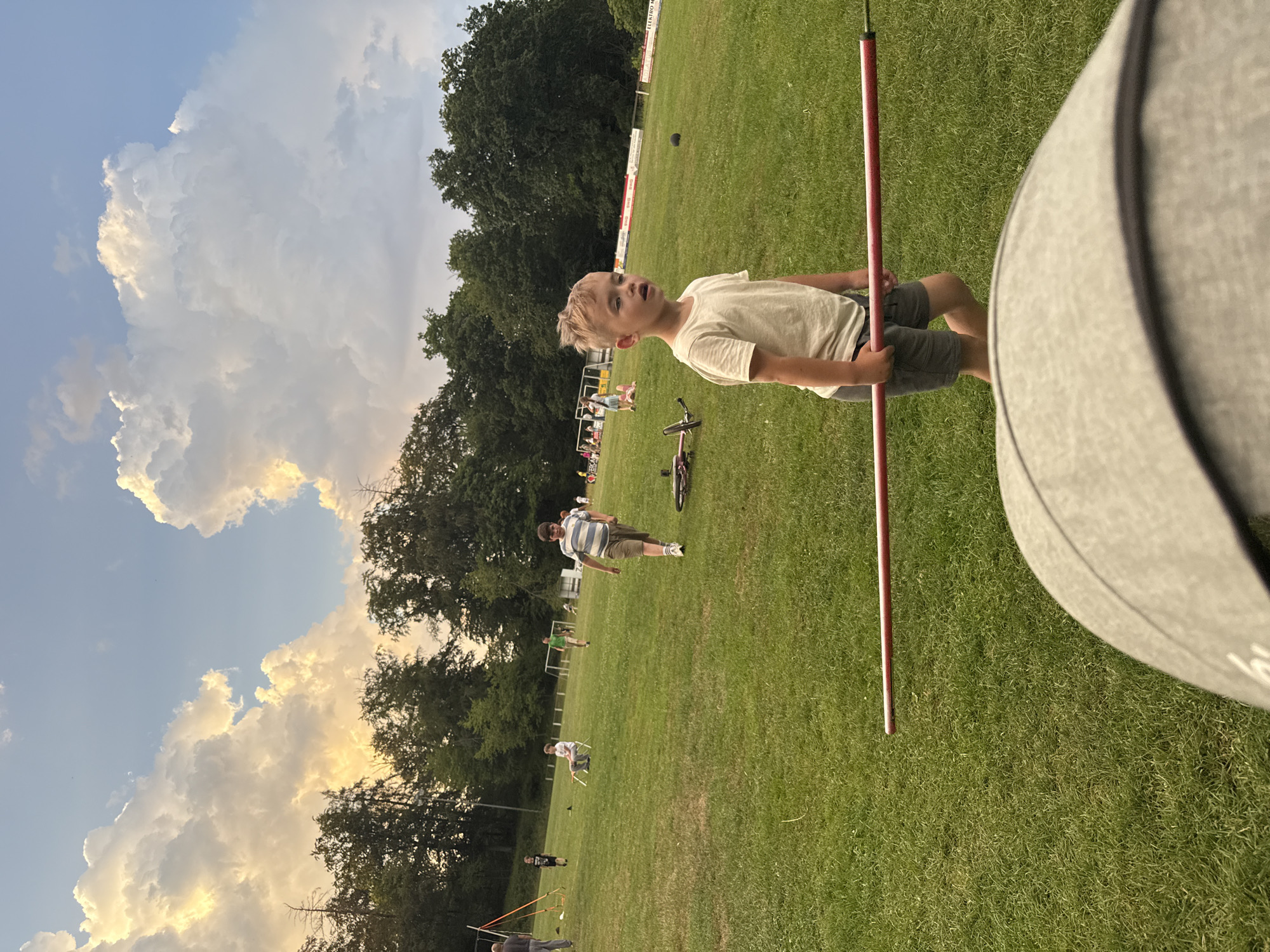 The image depicts a grassy outdoor field, likely a sports field, in Lumda, Germany. In the foreground, a young boy with blond hair is walking, holding a long red pole. He is wearing a light-colored t-shirt and gray shorts. Behind him, other children and adults are scattered across the field, some engaged in activities. One boy in a striped shirt is walking towards the foreground, while another sits on a chair in the middle ground. A bicycle is lying on the grass to the right of the boy holding the pole. The background features a line of trees and a cloudy sky, with the setting sun casting a warm glow on the clouds. There are also some sports equipment and structures visible, such as goalposts and banners. The overall scene suggests a casual outdoor gathering or event.