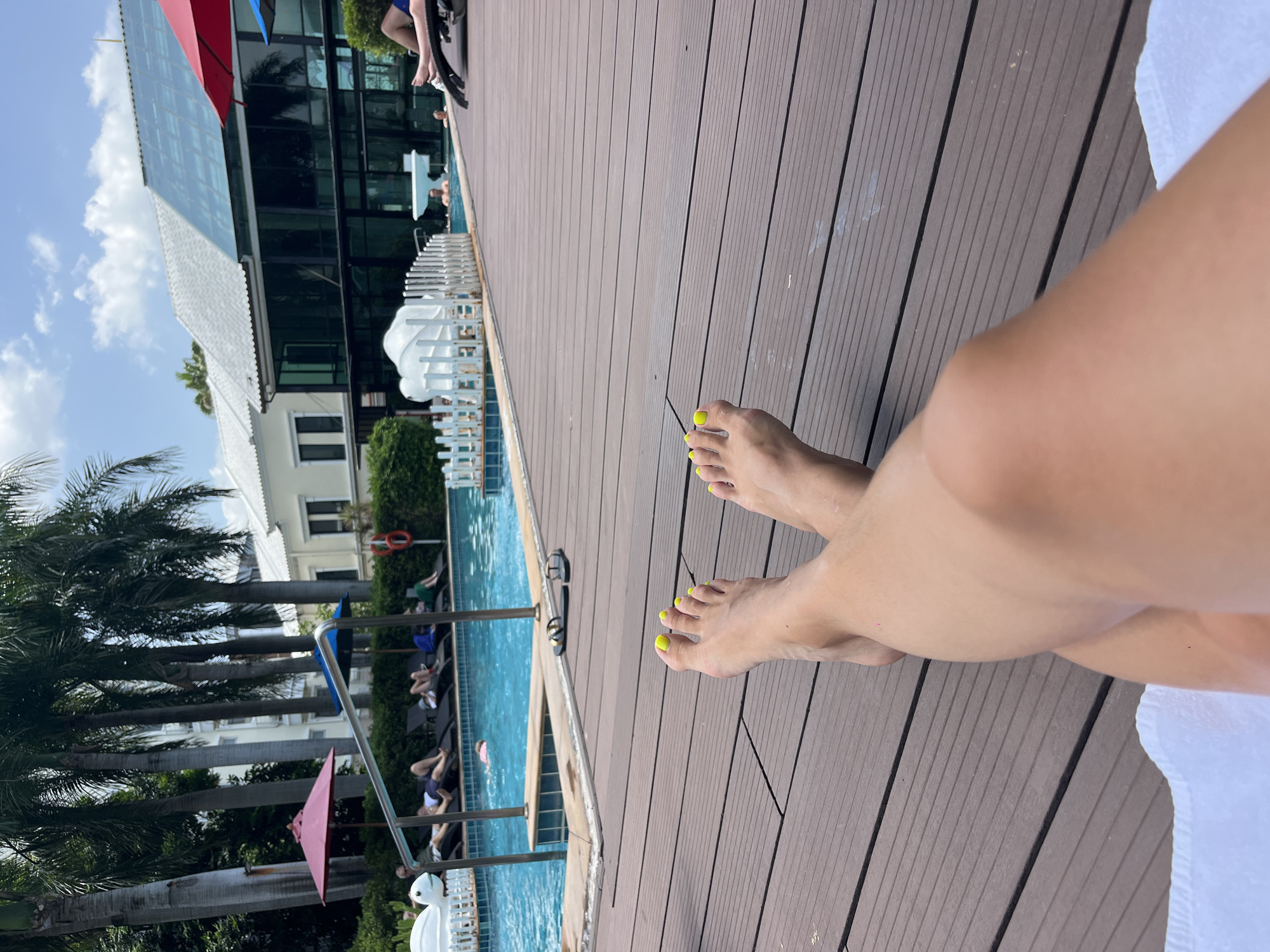 In Bang Sao Thong Subdistrict, Thailand, a person's legs, with bright yellow toenails, are prominently featured in the foreground, resting on a wooden deck by a swimming pool. In the background, the pool area is visible, with lounge chairs, umbrellas, and several people relaxing. Some individuals are in the pool, while others are lying on the chairs. The scene is set against a backdrop of tropical vegetation and modern buildings, under a partly cloudy sky.