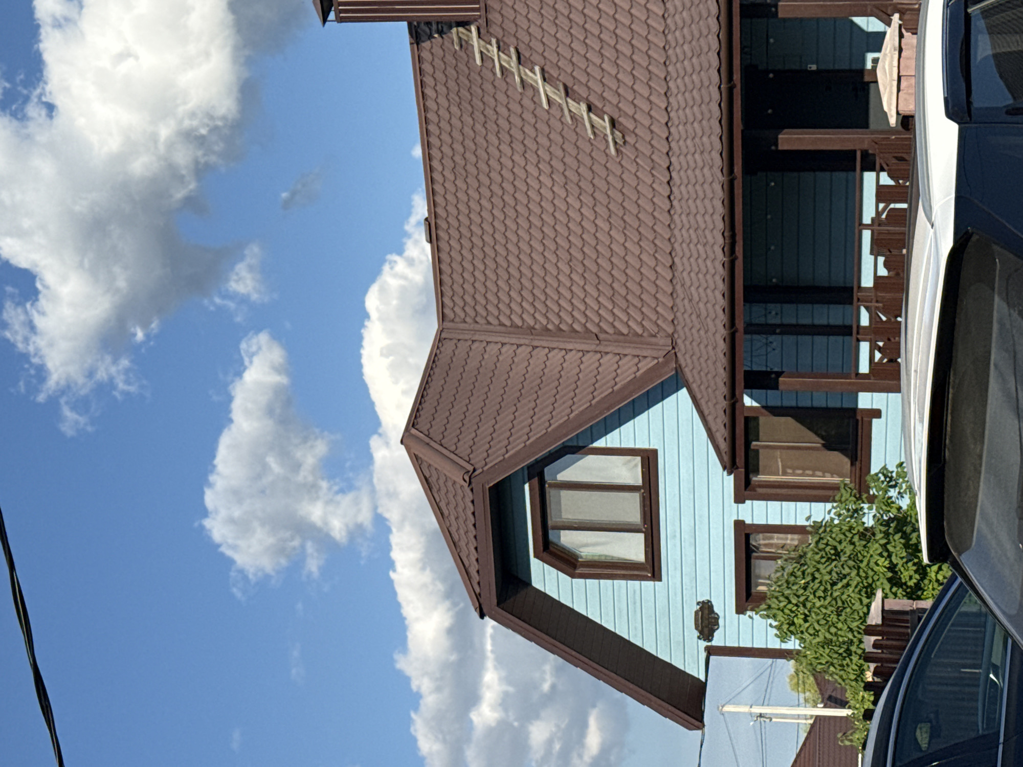 The image shows a light blue house with a brown roof in the village of Nagaevo, Russia. The house has a prominent gable roof with a small ladder attached to it. There are several windows visible on the house, some with brown frames. A porch with a wooden railing is present in front of the house. In the foreground, the top of two cars is visible, one white and one black, partially obscuring the lower portion of the house. Lush green foliage is growing in front of the house, behind a wooden fence. The sky is blue with fluffy white clouds. There are no people visible in the image. The scene appears to be a residential area on a sunny day.