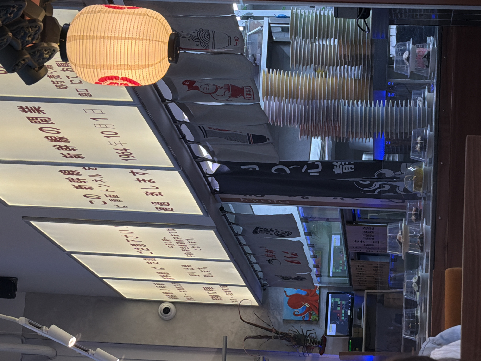 The image depicts the interior of a sushi restaurant, likely in Saint Petersburg, Russia, given the context of the provided crops. In the foreground, several plates of sushi covered with plastic domes are arranged on a conveyor belt system. Behind the conveyor belt, stacks of colorful plates are visible on shelves. The restaurant features Japanese-style decor, including hanging noren curtains with illustrations, a traditional lantern, and signage with Japanese text. A decorative lobster and a painting of an octopus are also visible on the left side of the image. In the background, a security camera is mounted on the wall. The lighting appears to be a mix of natural light filtering through illuminated panels and artificial lighting, creating a warm ambiance.