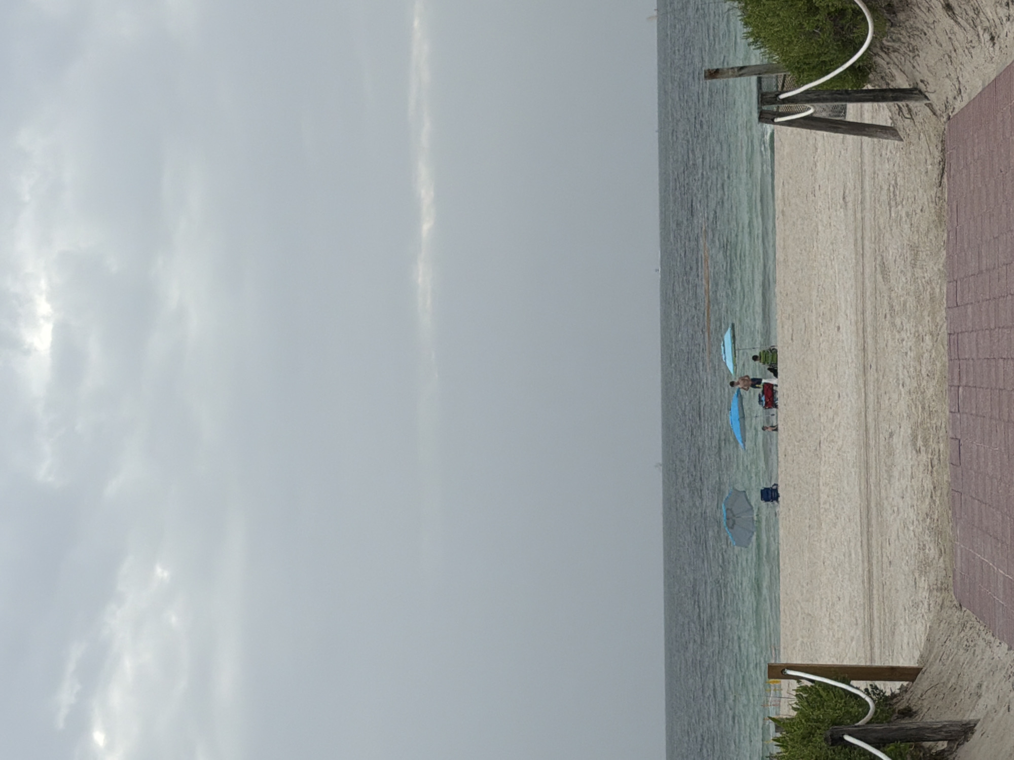 This is a vertical, eye-level, medium shot of a beach scene in Miami Beach, Florida, USA. The sky is overcast with a light gray hue, and there are some darker clouds visible in the upper portion of the frame. The ocean, a muted turquoise color, occupies the middle third of the image, with gentle waves rolling towards the shore.
On the sandy beach, there are three beach umbrellas providing shade. To the left, a light blue umbrella is open, and a person in a green chair is seated beneath it. In the center, a larger light blue umbrella is open, with a person standing nearby. To the right, another light blue umbrella is open, and a person in red shorts is standing next to a red cooler or beach cart. There are other beachgoers in the distance, but they are not clearly discernible.
The foreground of the image features a brick pathway leading towards the beach, bordered by sandy dunes with green foliage on either side. Wooden posts with white ropes delineate the pathway and the edge of the vegetation. The overall atmosphere is subdued, likely due to the overcast weather.