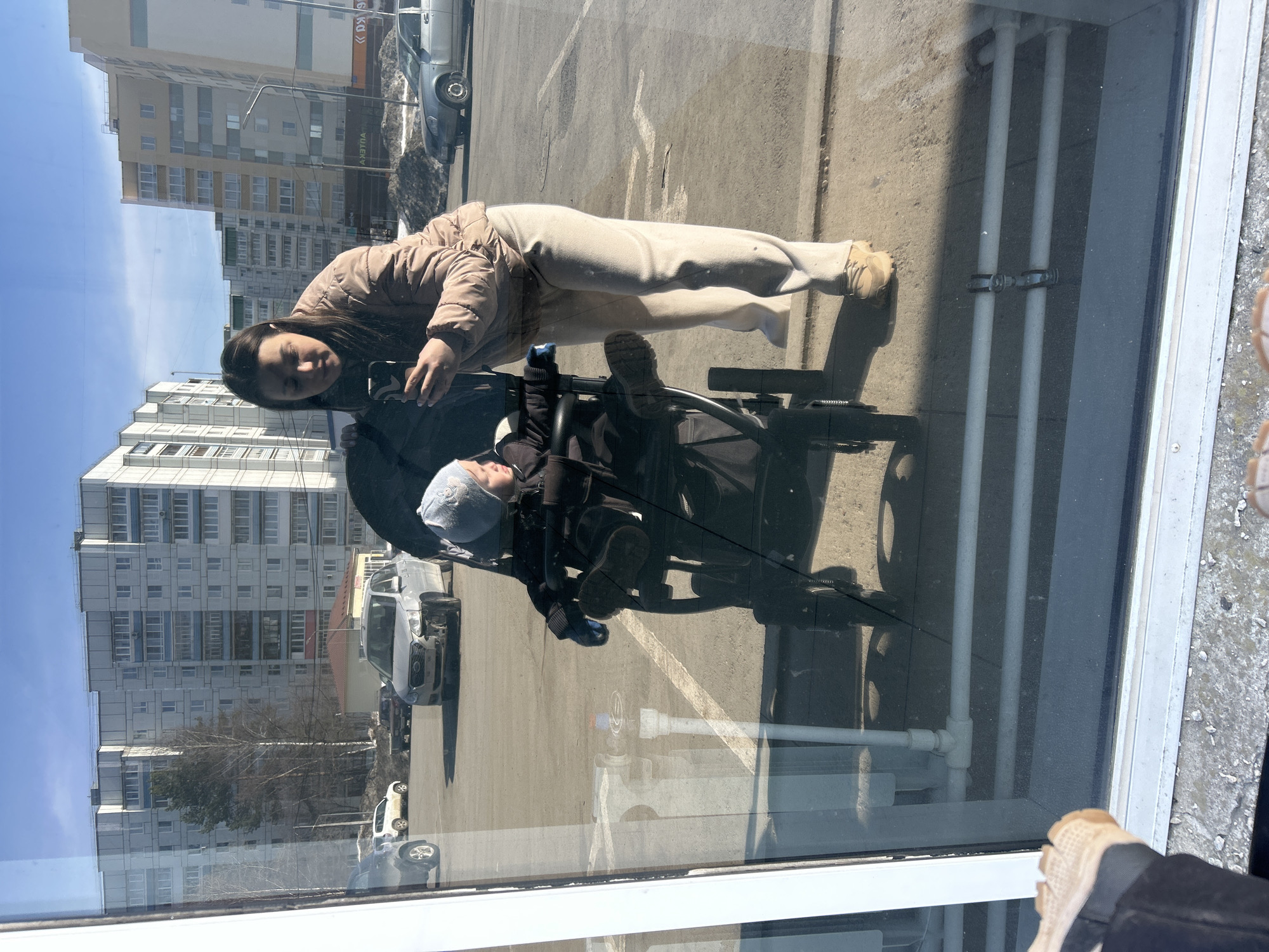 The image depicts a reflection in a window in what appears to be the city of Chaikovsky, Russia. A woman in a beige jacket and light-colored pants is holding a phone, possibly taking a picture. In front of her is a baby in a stroller. The baby is wearing a hat and dark clothing. The reflection shows a building with many windows, cars parked in a parking lot, and trees. The weather seems sunny. Parts of a window frame and pipes are visible, indicating that the image is a reflection seen through a window.
