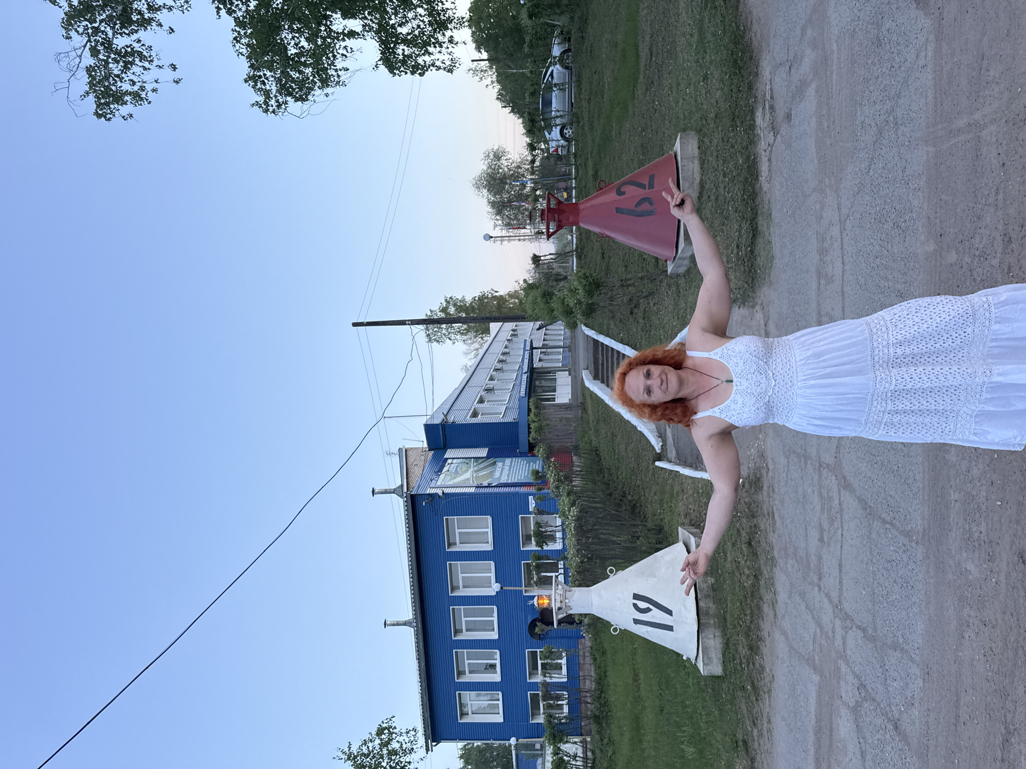 A medium outdoor shot depicts a fair-skinned, auburn-haired woman in a white dress, positioned on a concrete sidewalk alongside a building. She is holding two navigation buoys numbered "19" and "62". The building behind her is a three-story structure, predominantly blue, with multiple white-framed windows and a light fixture visible between windows. The location is Chaikovsky, Russia. The weather appears pleasant, indicated by the clear sky and light. In the background, trees, a utility pole, and parked cars can be observed. The woman is smiling slightly, and gives a peace sign above her head.