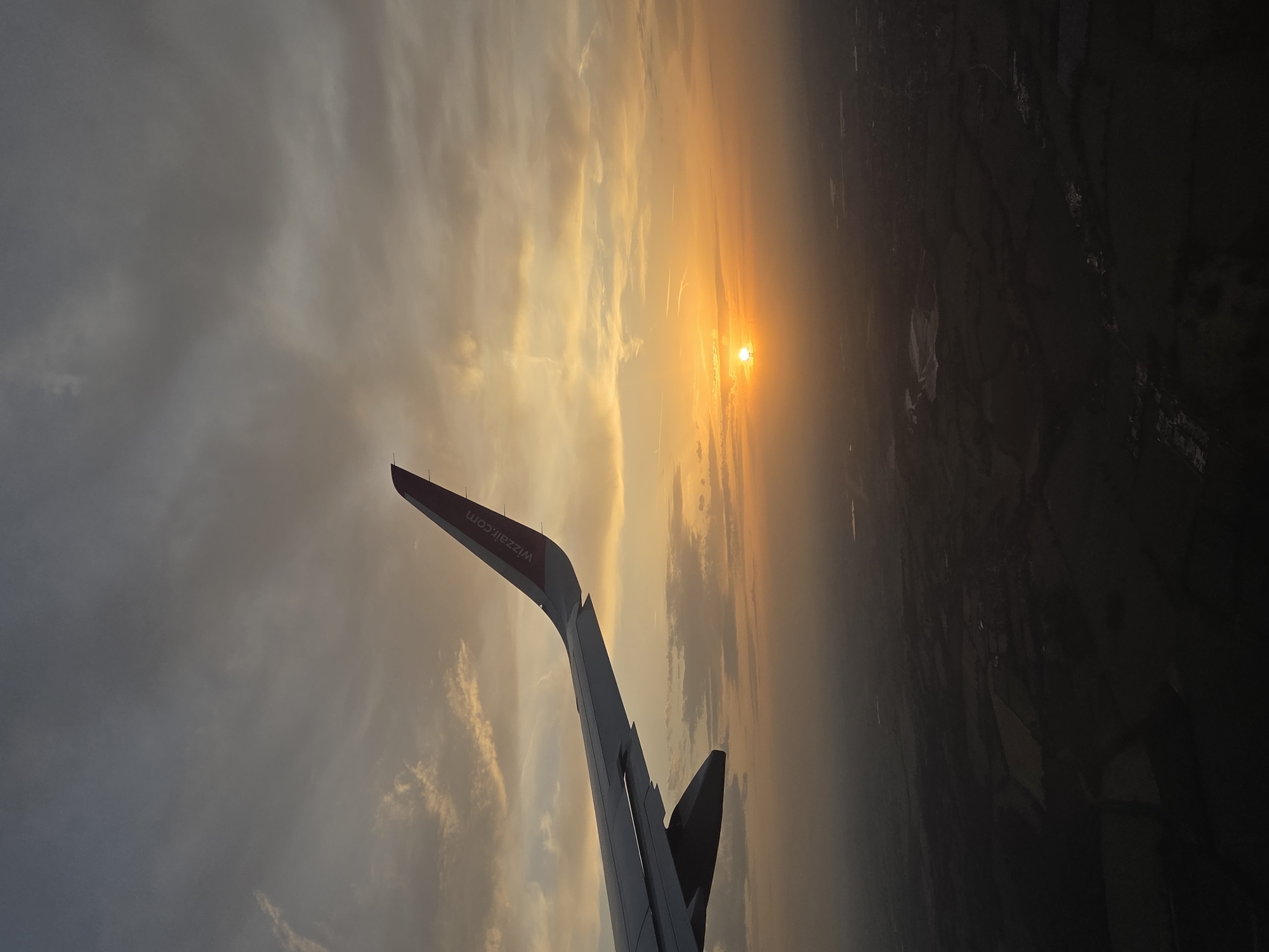 The image shows a view from an airplane window, likely during sunrise or sunset. The wing of the plane, identifiable by the "wizzair.com" marking, is visible in the foreground. The sun appears low on the horizon, obscured by atmospheric haze and clouds. Below the horizon, a dark, indistinct landscape is visible, possibly rural areas or a distant cityscape. Given the airline, this landscape is likely in or near the city of Dacorum, United Kingdom. There are no people visible, and the main subjects are the plane wing, the sky with clouds and sunlight, and the implied landscape below. The action is the flight itself, captured in a still image. The environment is the aerial perspective during a period of golden light.