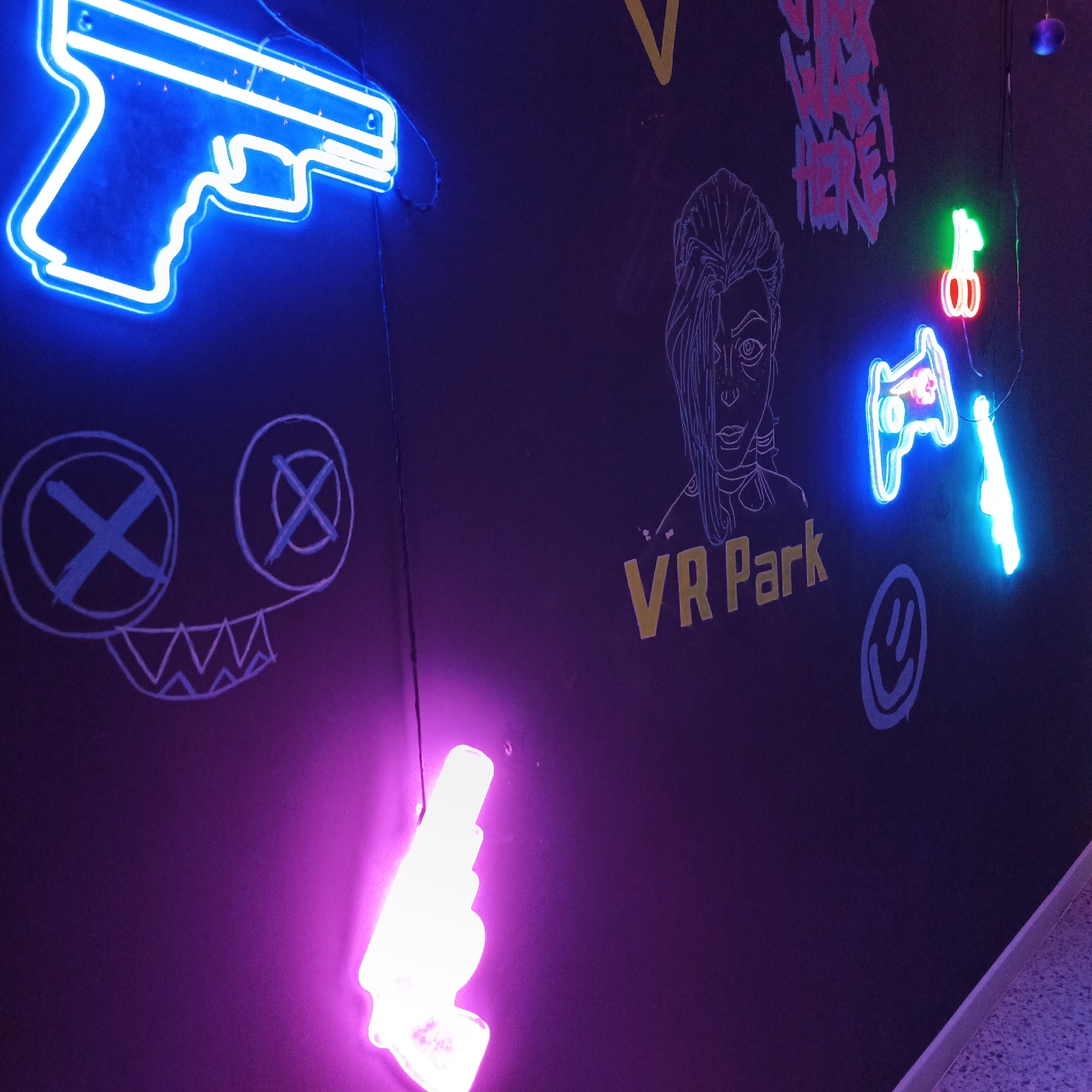 The image depicts an interior wall at "VR Park" in Chaikovsky, Russia. The dark wall features several neon signs, including the shape of a gun in blue, a pair of cherries in red and green, and another gun in cyan. There are also hand-drawn white chalk images of a face with crossed-out eyes and a jagged smile, a female character's face, and a smiley face. The words "VR Park" are painted in yellow near the center. There is a white gun-shaped neon sign near the bottom, bathed in purple light. There are various other painted writings. The floor is a speckled terrazzo-like material.