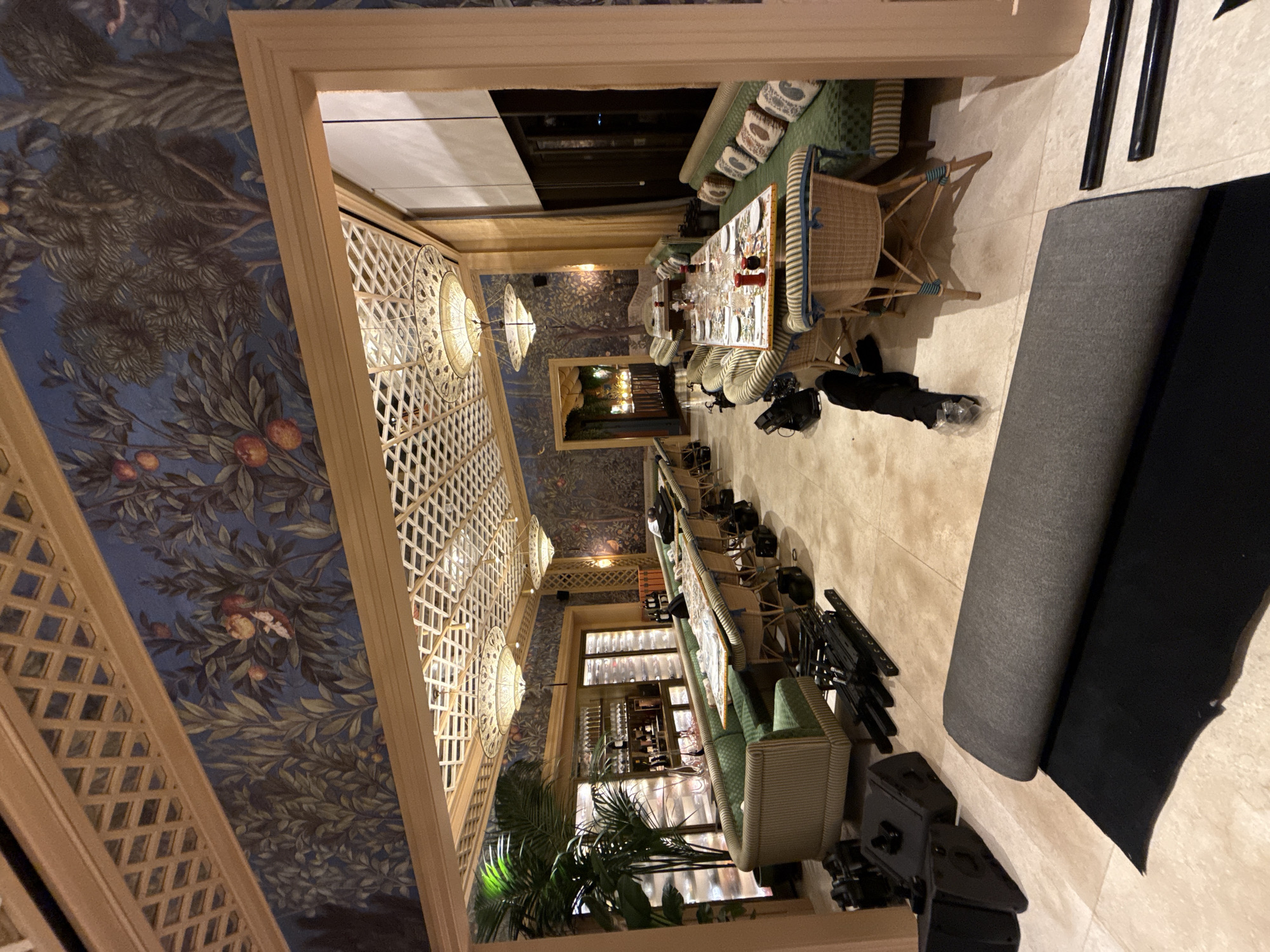 The image depicts the interior of a restaurant, possibly under renovation or setup, as evidenced by the rolled-up carpet in the foreground and various pieces of equipment on the floor. The restaurant features ornate wallpaper with a botanical motif of trees and fruit on a dark blue background. The ceiling has a decorative wooden lattice work and elaborate chandeliers.
Seating arrangements include curved banquettes upholstered in green striped fabric with patterned throw pillows, and individual wicker chairs. Several tables are set with placemats, tableware, and what appear to be salt and pepper shakers. A long bar area with bottles and glassware is visible in the background, as is a large plant in a pot.
Black audio speakers and metal stands are scattered on the tiled floor. The overall impression is of a sophisticated dining establishment with a distinct decorative theme. The prompt specifies the location as Baku, Azerbaijan, however, there are no visible elements in the image that definitively confirm this specific city.