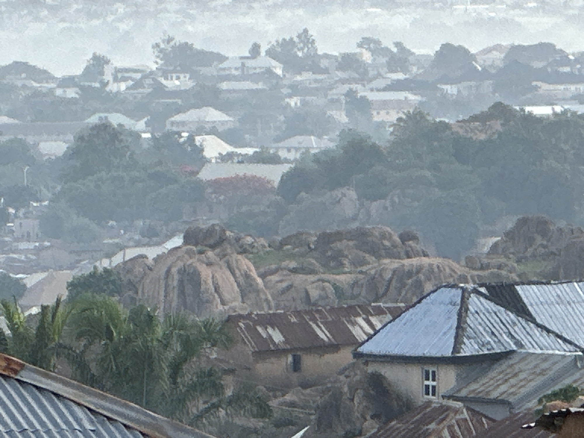 The image shows a hazy view of Jos, Nigeria. In the foreground are rooftops constructed of corrugated metal, some rusty and some painted silver, indicating varying levels of upkeep. Interspersed among the roofs are palm trees and weathered, beige-colored buildings. Behind these, large, rounded rock formations rise, partially obscured by vegetation. In the distance, more buildings are visible, with faint hints of red roofs. The atmosphere is overcast, with a layer of haze or fog reducing visibility and muting colors. The scene suggests a hilly or elevated terrain, with structures built amidst the natural rocky landscape.