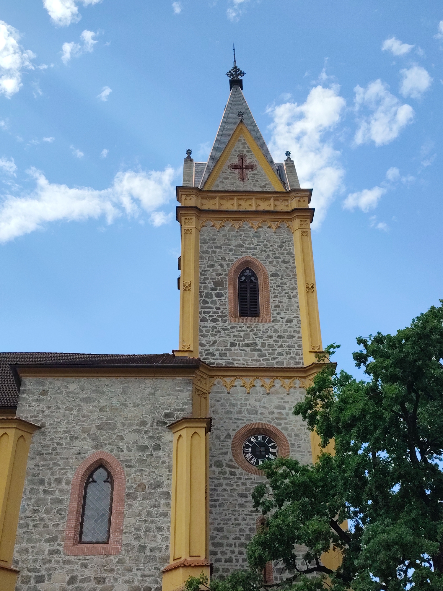 The image shows the tower of a stone church, painted in yellow accents, under a bright blue sky with scattered white clouds. The church has a pointed roof topped with a spire and ornamental crosses. A prominent clock face with Roman numerals is visible on the tower. A large green tree partially obscures the right side of the church. This is the Saint John of Nepomuk Church in Hluboká nad Vltavou, Czechia. There are no people or animals visible in the image.