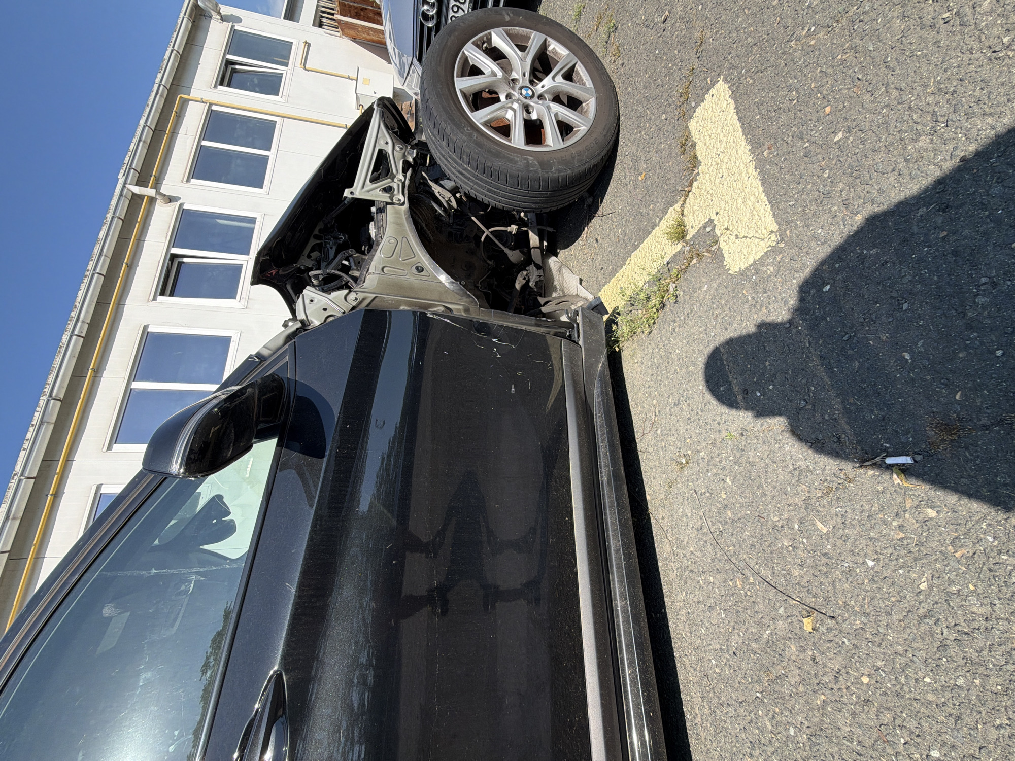 The image depicts a black vehicle that has sustained significant front-end damage. The front passenger-side wheel is heavily damaged and positioned at an unnatural angle. The car's front quarter panel is missing or severely deformed, revealing the vehicle's internal structure. The car is parked next to a building with multiple windows, likely an office or residential structure. The ground is asphalt and marked with a faded yellow directional arrow. A shadow, likely from the photographer, is visible on the pavement. Another vehicle is partially visible in the background. A cigarette butt is on the ground. It is not possible to determine the specific location within Burgas, Bulgaria from this image alone. No people are visible.