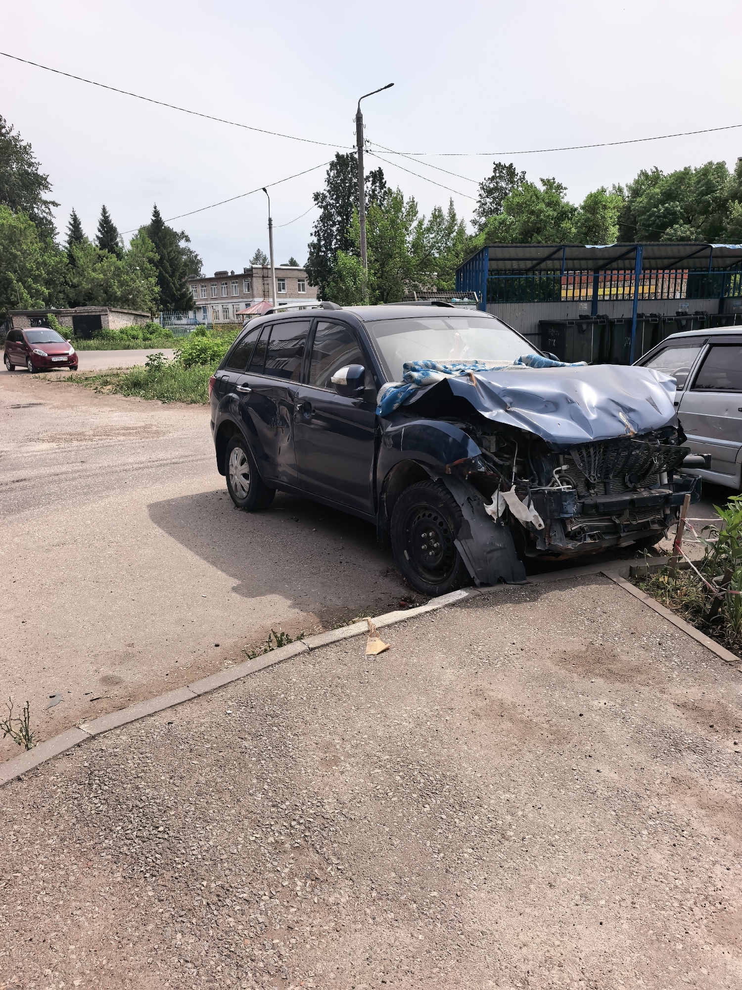The image shows a dark blue car with severe front-end damage, including a crumpled hood, broken front bumper, and exposed engine components. The car is parked on a gravel or paved area near a curb. The scene appears to be in a residential or commercial area in Sterlitamak, Russia, suggested by the presence of buildings and other cars in the background. There is a small red car parked on the street in the background to the left. A dumpster enclosure and a silver-colored car are present in the background on the right. The sky is overcast. There are no people visible in the immediate vicinity of the damaged car.