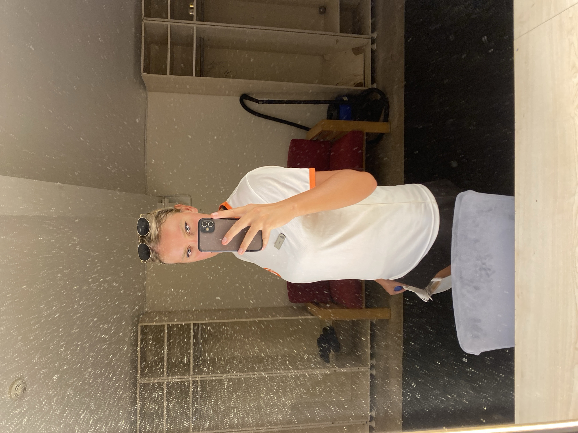 In a room with multiple open lockers, a young woman with blonde hair tied up, wearing sunglasses on her head and a white polo shirt with orange trim, is taking a mirror selfie. She is holding a smartphone in her right hand and her left hand is near her lower body. In the background, a red chair and a blue vacuum cleaner are visible. The room appears to be a locker room or changing area. The overall lighting is dim, and the mirror surface is speckled with dust or water droplets.
