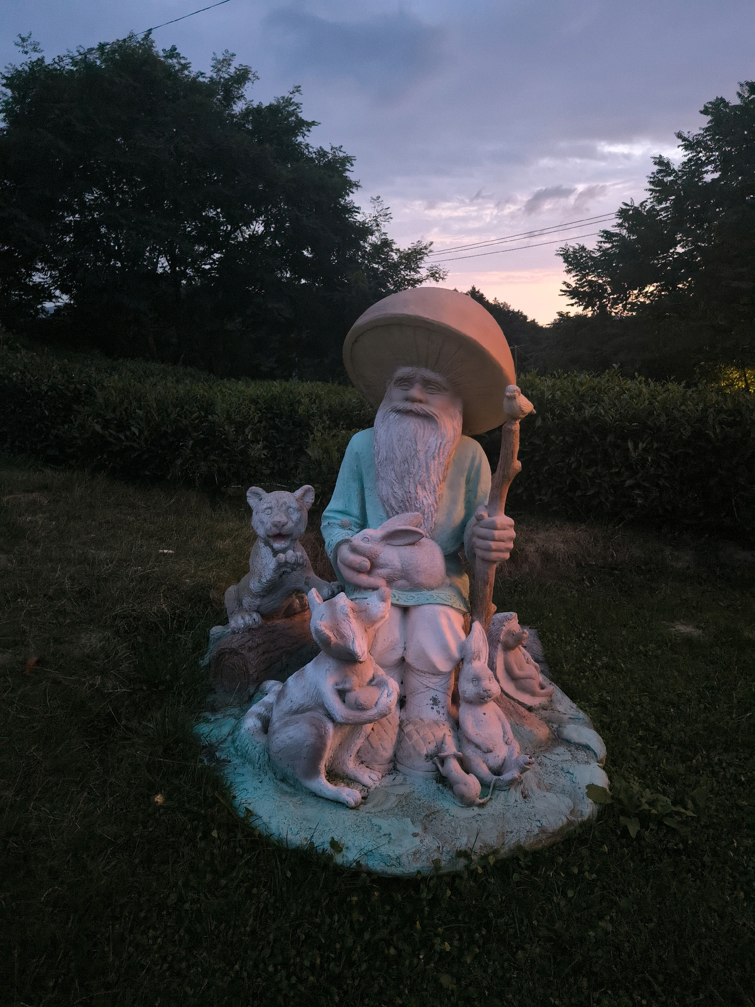 This is a photograph taken at dusk in Kalinovoye Ozero, Russia. The focal point of the image is a garden gnome statue. The gnome is depicted as an elderly man with a long white beard and a blue tunic, holding a staff. He is seated and surrounded by several animal figurines, including rabbits, a squirrel, and a small cat-like creature. The gnome and animals are resting on a turquoise-colored base, which is placed on a grassy area. Behind the statue, there is a dense hedge and a line of trees. The sky above is a twilight gradient of blue and purple, with hints of pink from the setting sun. A power line is visible stretching across the sky. The lighting suggests it is either early morning or late evening.