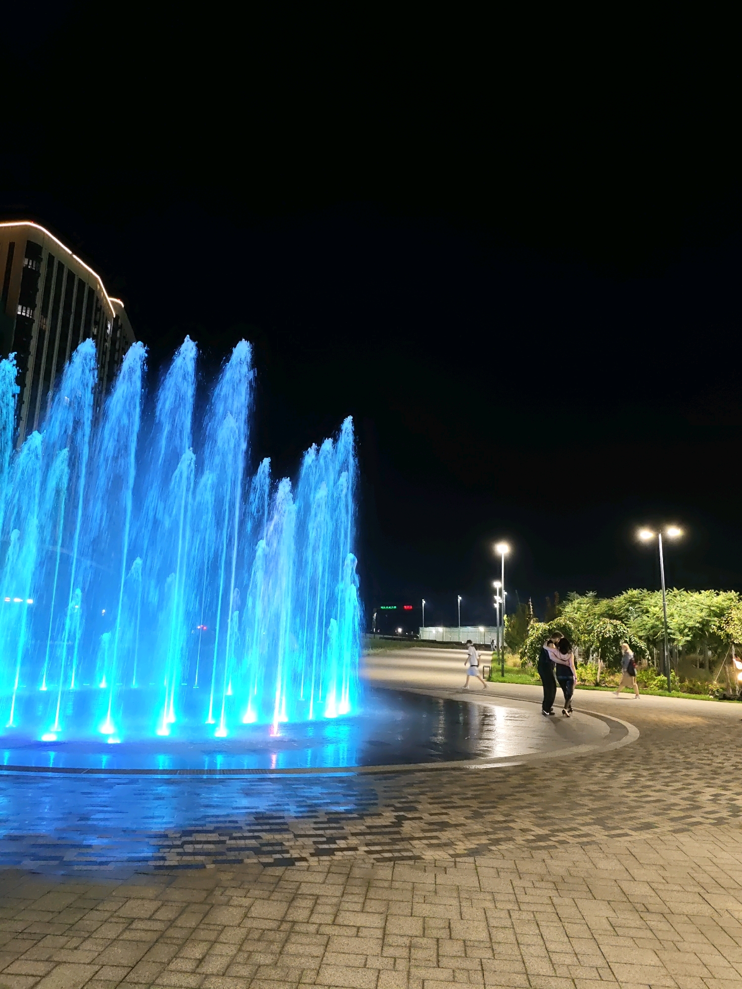 A nighttime view of a contemporary urban plaza features a large, illuminated fountain with vibrant blue water jets. To the left, a modern building with glowing accents stands against the dark sky. The plaza's ground is paved with interlocking bricks, reflecting the fountain's light. Several people are visible walking on the paved path to the right of the fountain, with a couple embracing in the mid-ground. Streetlights illuminate the area, casting a warm glow on the surroundings. The scene is set in the city of Rostov-on-Don, Russia.