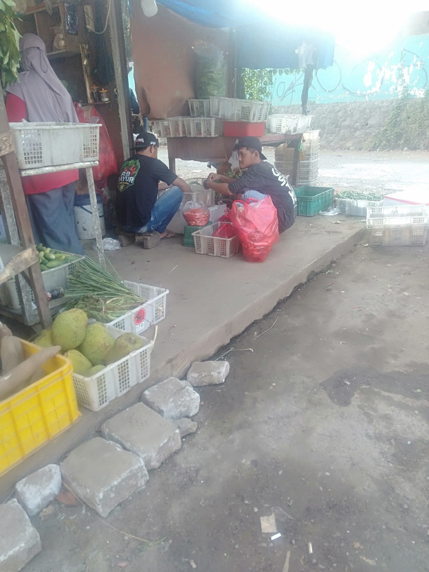 At a roadside produce stand in Sukadami, Indonesia, a vendor wearing a light purple hijab and a red shirt stands behind a white crate filled with cucumbers. To her right, two young men are seated on the ground. One, wearing a black t-shirt with white lettering that reads "AYUR Bahagia," is sorting through red chilies in a white plastic basket. The other, in a dark t-shirt, is arranging items in a large red plastic bag. Various baskets containing produce, including jackfruit and long green stalks of what appear to be lemongrass, are visible. In the foreground, a bright yellow crate is positioned alongside other produce. The ground is a mix of concrete and dirt, with several large stones scattered around. The background shows a stone wall with teal graffiti and a blue tarp overhead, typical of a street-side market.