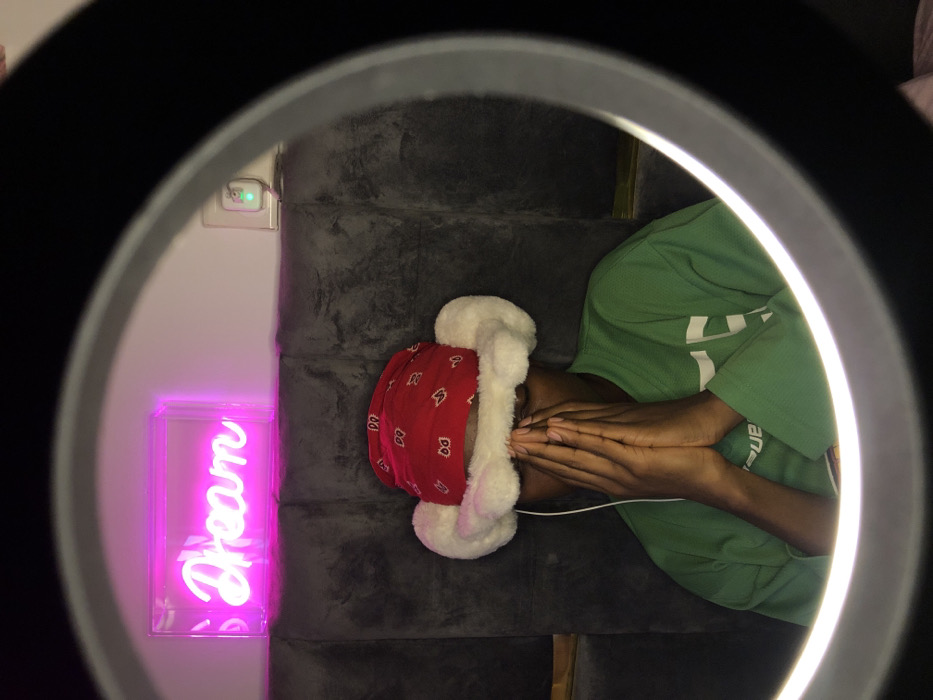 An individual is depicted centrally, framed by the illuminated ring of a black ring light in the foreground. The person wears a red bandana with white patterns beneath a white, fluffy, ear-flap style hat, and a long-sleeved green shirt. Their hands are clasped together, obscuring their face from the nose downward. A white earbud cable is visible, extending from the subject's left ear area. Behind the individual, a dark grey, padded, tufted headboard or wall panel serves as a backdrop. Above this panel, mounted on a white wall, a pink neon sign spelling "Dream" in cursive script is illuminated. To the upper right of the neon sign, a white power outlet with an active green indicator light is visible, indicating a plugged-in device. The scene is illuminated by the ring light and the neon sign, suggesting an indoor setting. This scene is located within the city of Tema, Ghana.