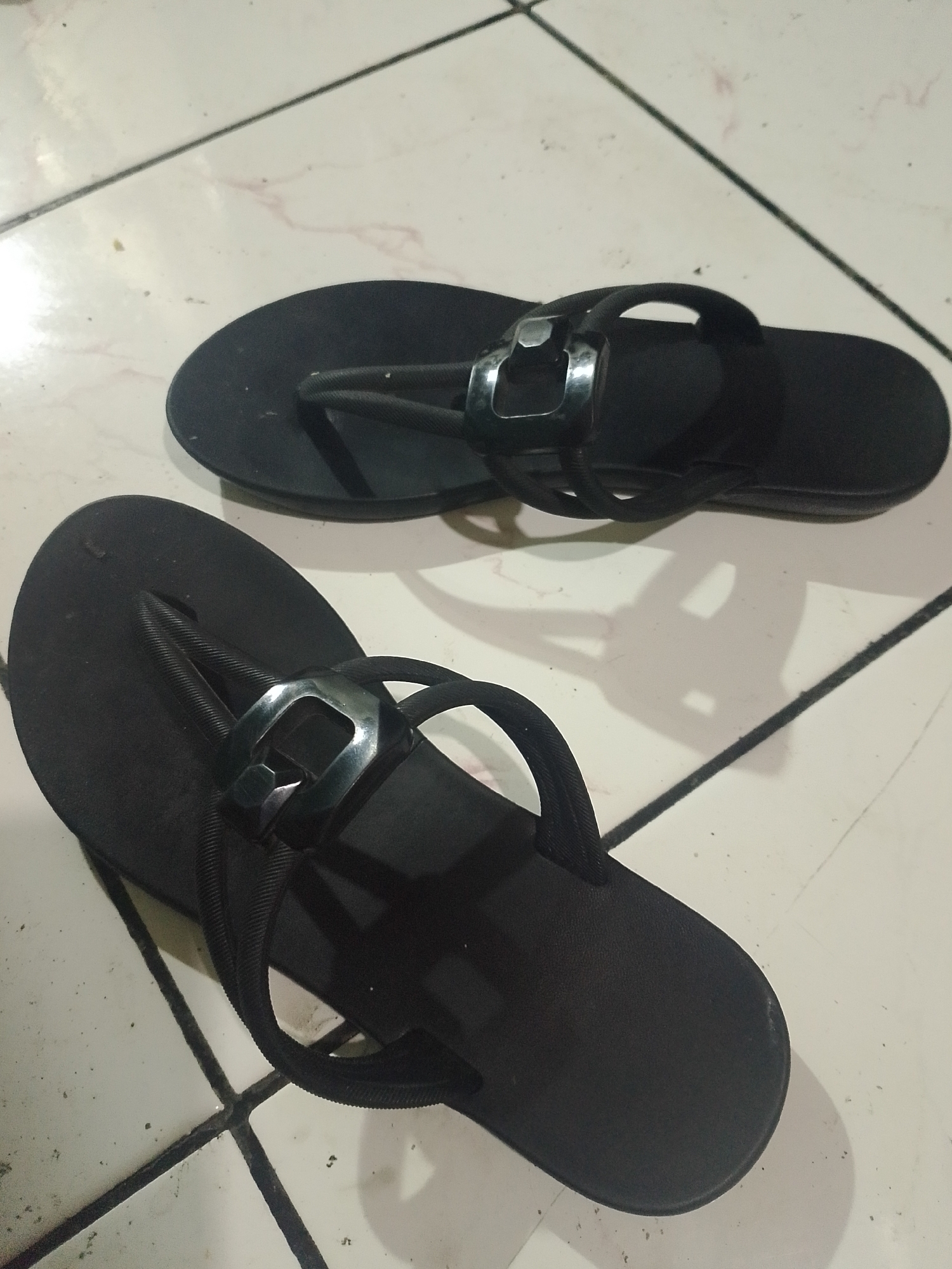 A pair of black flip-flops are displayed on a tiled floor in Mukapayung, Indonesia. The flip-flops are positioned with the straps facing upward, revealing the soles. Each flip-flop features a black strap that goes between the toes and then splits into two straps that wrap around the sides of the foot, secured by a metallic accent piece. The tiled floor has a light color with dark grout lines. The lighting in the scene suggests an indoor setting, possibly a home. There are no people present in the image.