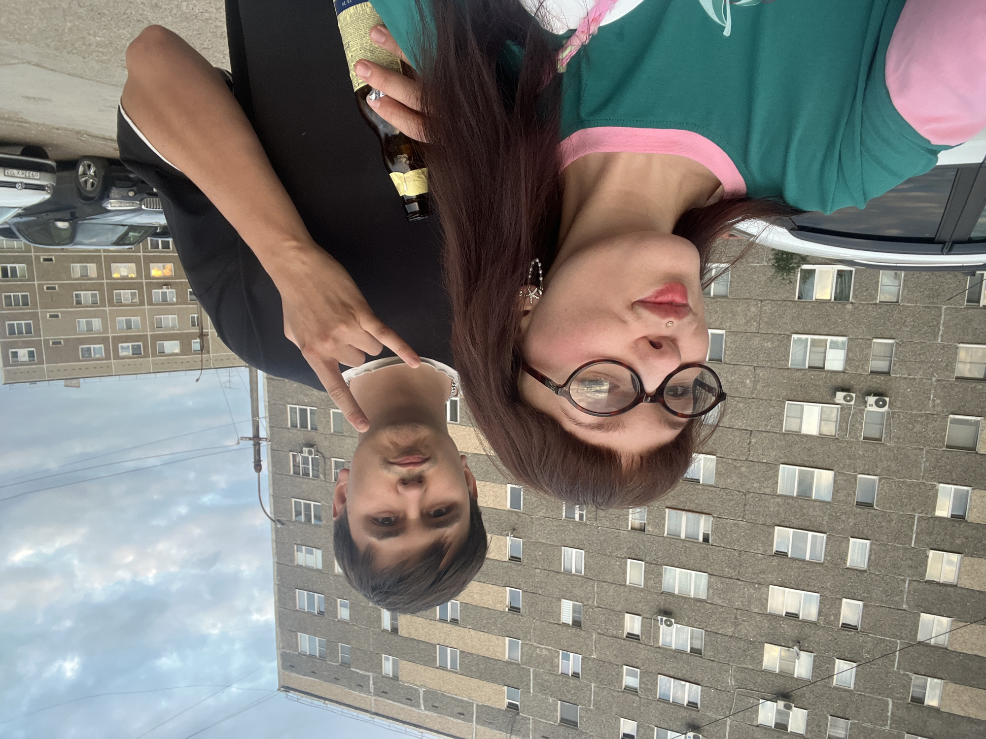 In Chaikovsky, Russia, a young woman with brown hair, glasses, and a septum piercing poses for a selfie with a man. She wears a green and pink long-sleeved shirt and is holding a beer bottle. The man, with dark hair, wears a black t-shirt and makes a rock-on hand gesture. Behind them, a large, grey, Brutalist-style apartment building stands under a cloudy sky. Several cars are parked in the foreground.