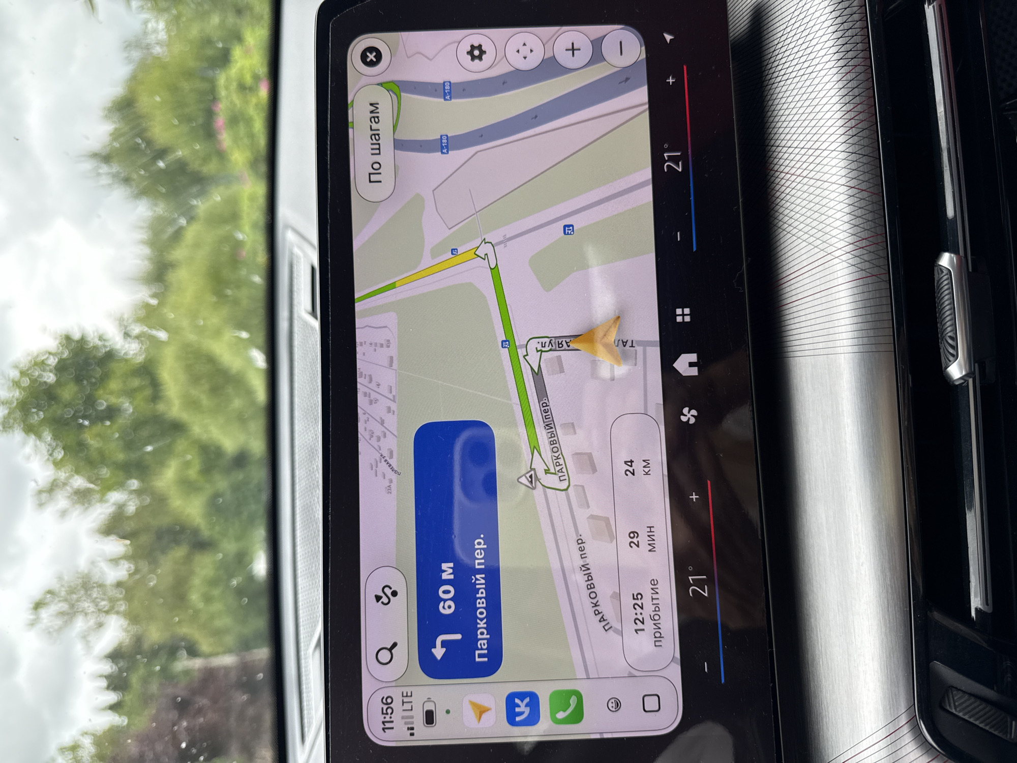 The image shows a navigation screen in a car, displaying a map with a route highlighted. The current location is indicated by a yellow marker. The screen shows the distance to the destination as 60 meters, with the street name "Парковый пер." (Parkovy per.) indicating the destination. It also shows an estimated arrival time of 12:25, with 29 minutes and 24 kilometers remaining. The current temperature displayed is 21 degrees Celsius. The overall scene is indoors, within a vehicle, with a view of trees and sky through the windshield. There is no indication of the specific location within Russko-Vysotskoye Sel'skoye Poseleniye, Russia, beyond what is shown on the navigation screen. There are no people visible in the image.
