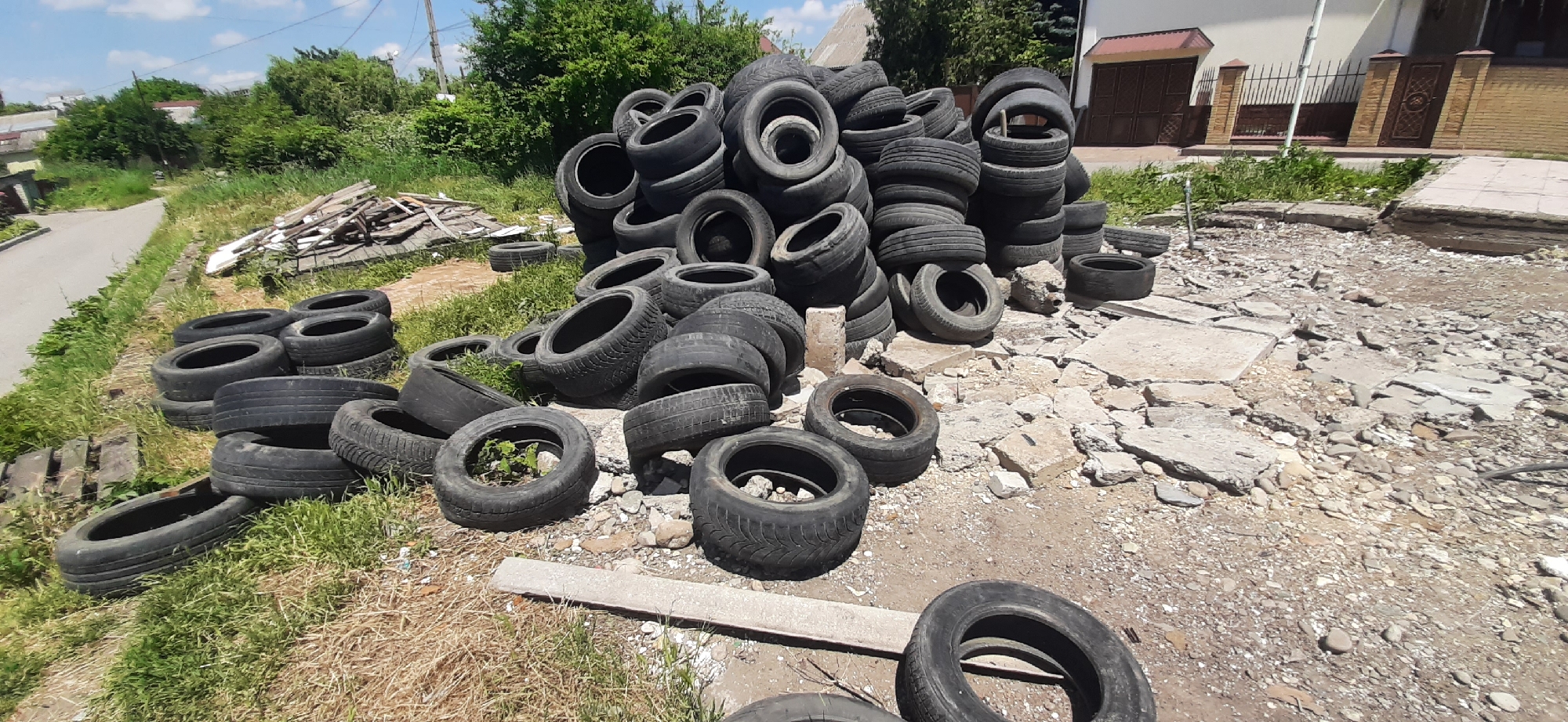 The image depicts an outdoor scene in Yessentukskaya, Russia, characterized by a large pile of discarded tires. The tires are of various sizes and conditions, some stacked vertically and others lying horizontally, forming a dense accumulation. They are scattered across a rough, uneven terrain composed of dirt, gravel, and broken concrete fragments. In the background, a paved road is visible on the left, bordered by overgrown grass and vegetation. Further back, there are buildings, including a residential house with a light-colored facade and a dark-colored roof, and a fence made of brick and metal. The sky above is blue with scattered clouds. No people are present in the image, and no specific actions or interactions are taking place. The overall scene suggests an informal dumping area or a site undergoing demolition or renovation.
