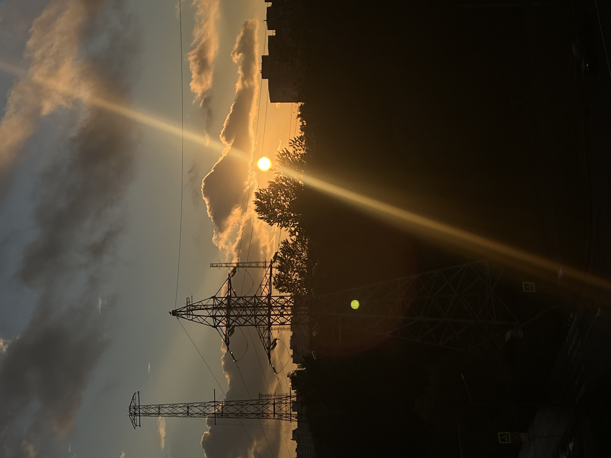 The image captures a sunset in Saint Petersburg, Russia, with a strong sunbeam cutting across the frame from left to right. The sky is a mixture of warm orange and yellow hues from the setting sun, interspersed with dark and light grey clouds. The sun itself is a bright, glowing orb visible behind a line of trees.
Dominating the foreground are several tall, lattice-like electricity pylons, silhouetted against the bright sky and the darkening landscape. The pylons are intricately structured with cross-bracing and support cables. The scene transitions into a dark, silhouetted area on the right, where the outlines of buildings and more trees are barely discernible. The lower part of the image shows a street-level view with what appear to be crosswalk signs. The overall mood is dramatic and atmospheric, emphasizing the stark contrast between the illuminated sky and the darkening foreground. The time of day is clearly sunset.