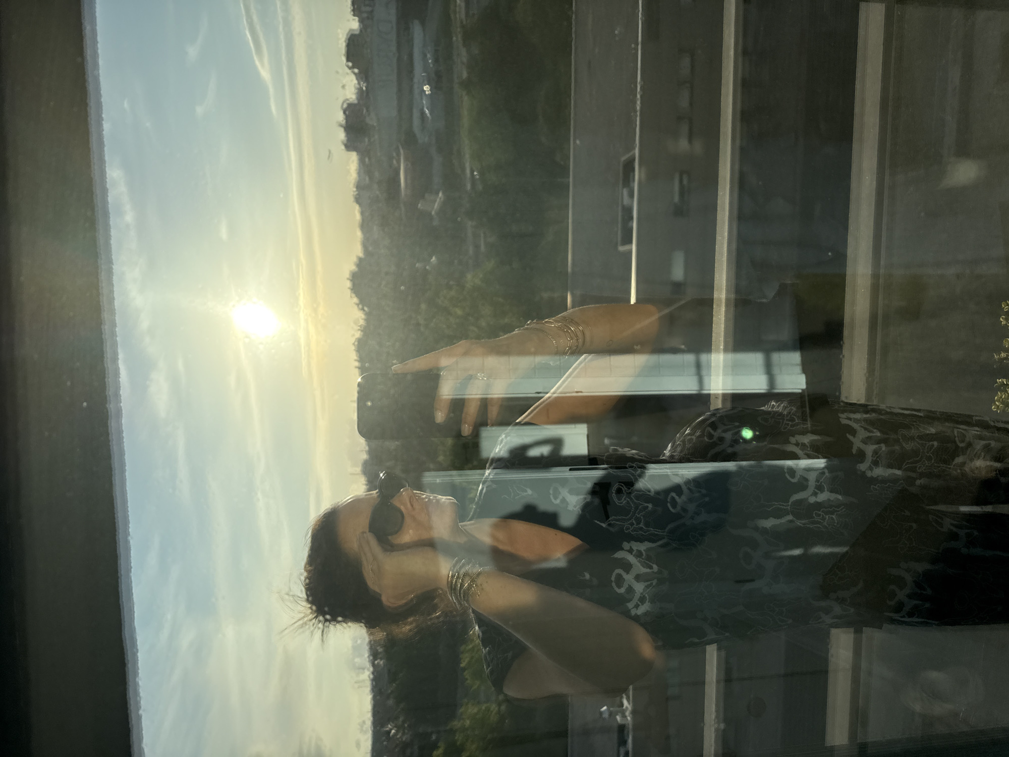 The media file captures a scene viewed through a window, featuring a person reflected or seen through the glass against a bright outdoor landscape. The image is taken in portrait orientation but depicts a horizontal scene, appearing rotated 90 degrees counter-clockwise.
The main subject is a woman with dark hair, wearing sunglasses and a dark patterned top or dress. She has multiple golden bracelets on both wrists. Her reflection is superimposed on the exterior view. One hand is raised, with her index finger pointing towards the bright sun in the distance, while her head is tilted slightly upwards, looking towards the horizon. Her other arm is bent, resting near her chest.
Outside the window, a bright sun is visible low in the sky, suggesting either sunrise or sunset. The sky is a clear, light blue with some subtle wispy clouds. Below the horizon, a sprawling urban or suburban landscape unfolds, featuring numerous buildings and what appears to be a multi-lane highway or major road with vehicles. Green trees and foliage are also visible, particularly on the left side of the frame. Vertical window frames or mullions divide the view, and reflections of the interior space are also visible on the glass.
The setting appears to be a high vantage point, possibly from a building or a vehicle. The time of day is clearly marked by the low, brilliant sun. No visible text is present in the image. The location context provided is Loures, Portugal, but the image itself does not contain specific geographical markers to confirm this.
