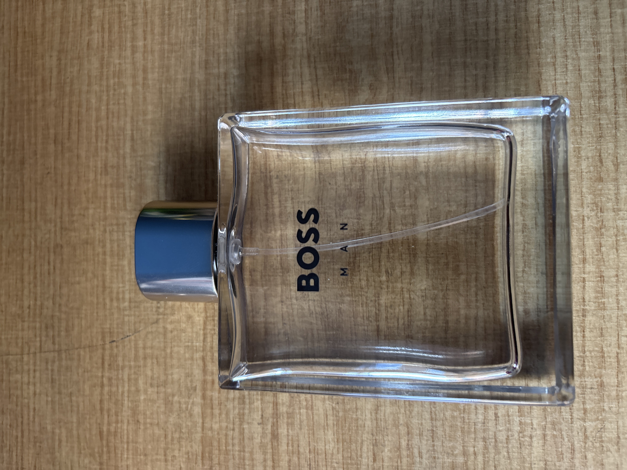 A clear glass perfume bottle is depicted lying horizontally on a light brown wooden surface. The rectangular bottle features slightly rounded edges and a prominent metallic silver-colored cap on its left end. A thin, transparent spray tube is visible inside the bottle, extending from the cap. Vertically printed in black capital letters on the bottle's face is "BOSS", with "MAN" horizontally printed in smaller black capital letters below it. The wooden background displays a distinct vertical grain pattern and variations in color, suggesting a natural wood texture. The image is a still life, likely taken indoors in Blagoevgrad, Bulgaria.