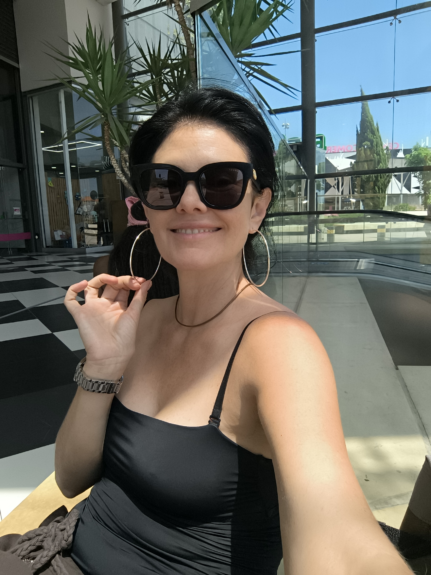 A woman with dark hair and sunglasses is taking a selfie. She is wearing a black dress and large hoop earrings. She is indoors, in what appears to be a shopping mall with a checkered floor and large glass windows. The environment suggests a modern architectural style. The setting is identified as the Alegro Setúbal shopping center in Setúbal, Portugal, indicated by reflected signage.