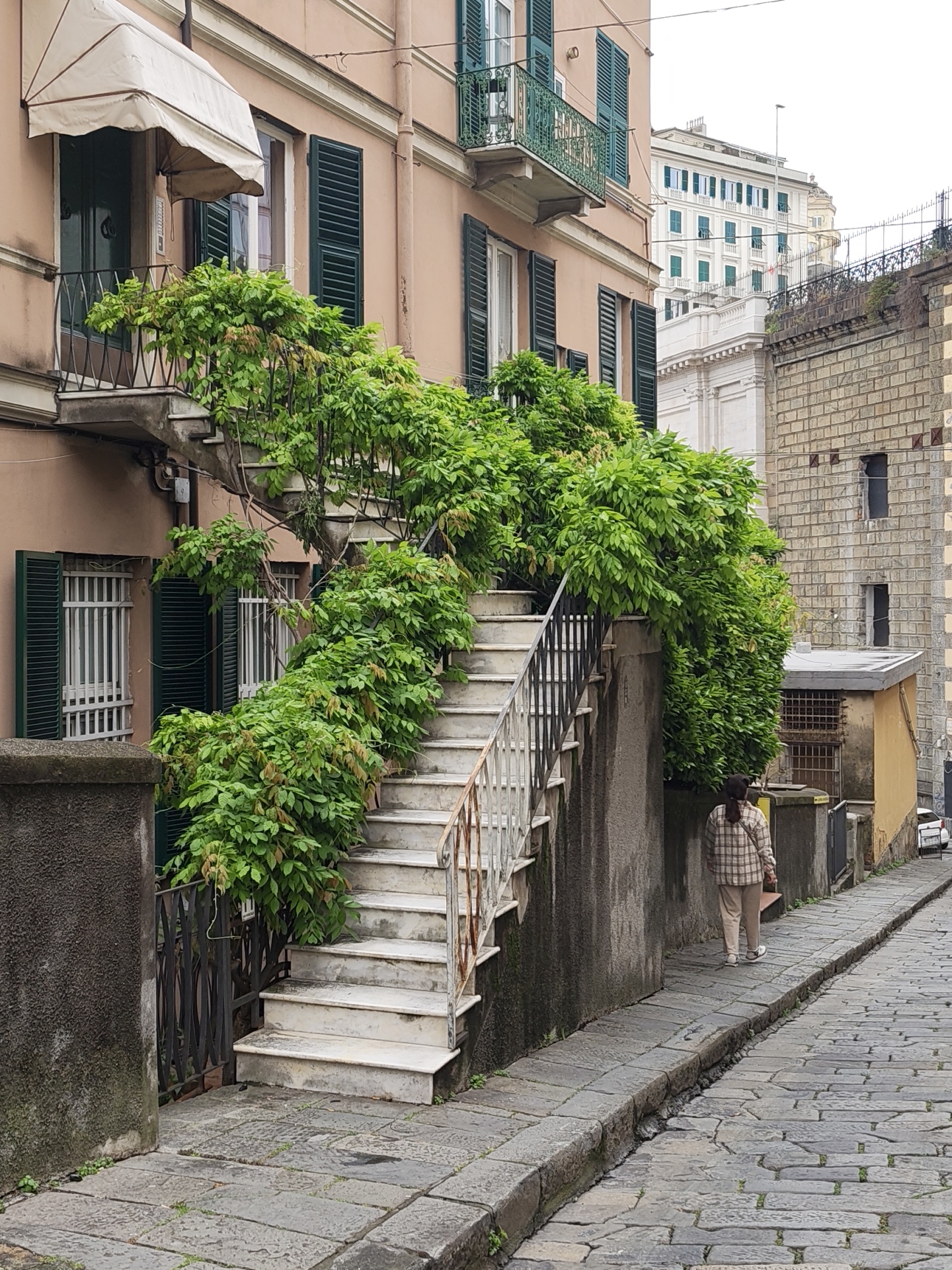 The image shows a narrow, cobblestone street in Genoa, Italy. The street ascends slightly. On the left is a peach-colored building with dark green shutters and a beige awning over a door on the second floor. An outdoor staircase, partially covered in lush green foliage, climbs from the street to the second floor. The staircase has a weathered white railing. A person wearing a plaid shirt and beige pants is walking away from the viewer along the right side of the street. The background features other buildings, including one made of tan brick and another taller white building with green shutters. The sky is overcast.