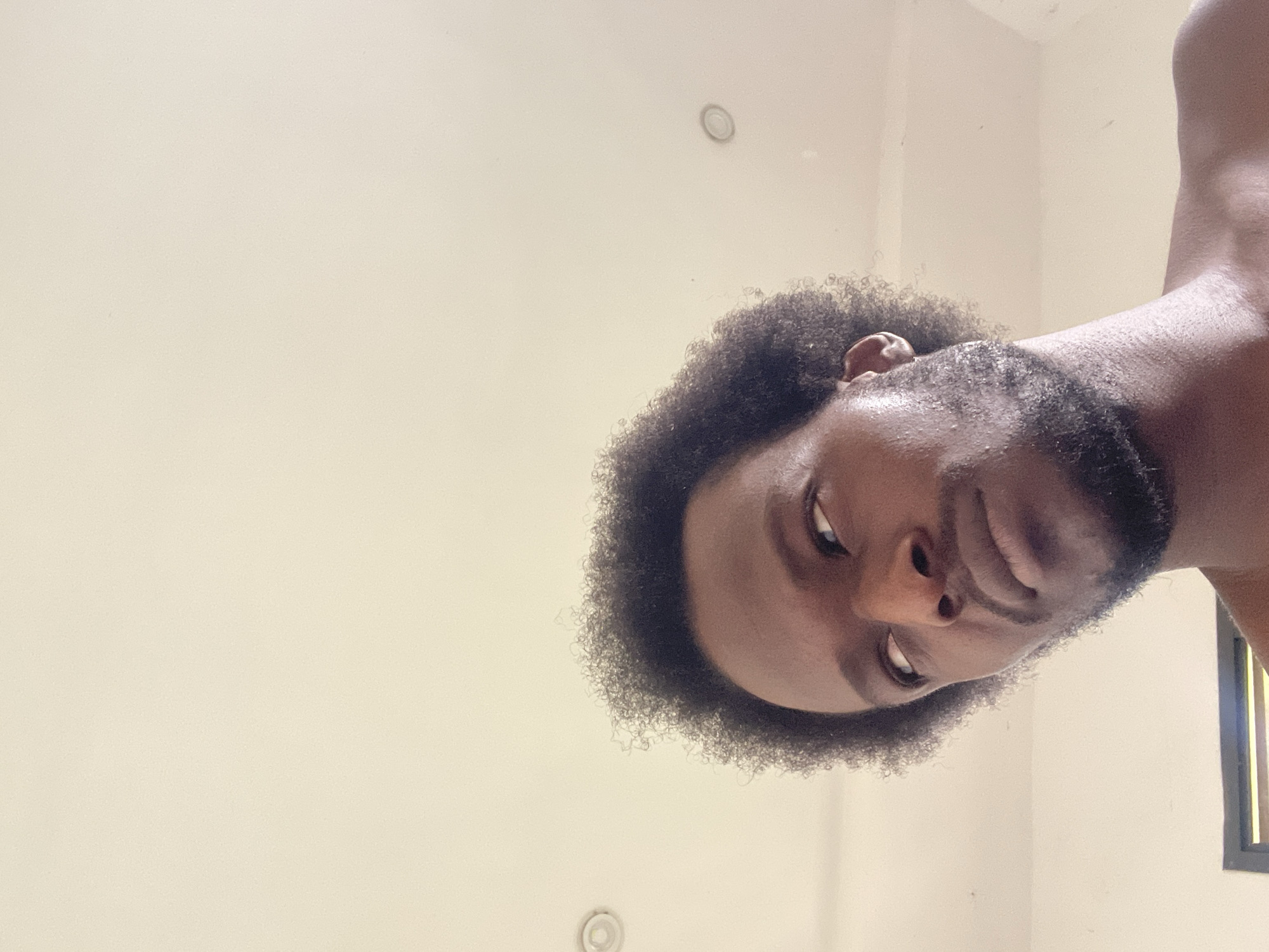 This is a close-up, low-angle shot of a Black man looking upwards and to his left. He has dark, curly hair and a beard. The background is a plain, light-colored ceiling with two circular light fixtures visible. The lighting suggests an indoor setting, possibly a home or hotel room in Abuja, Nigeria. The man appears to be shirtless.