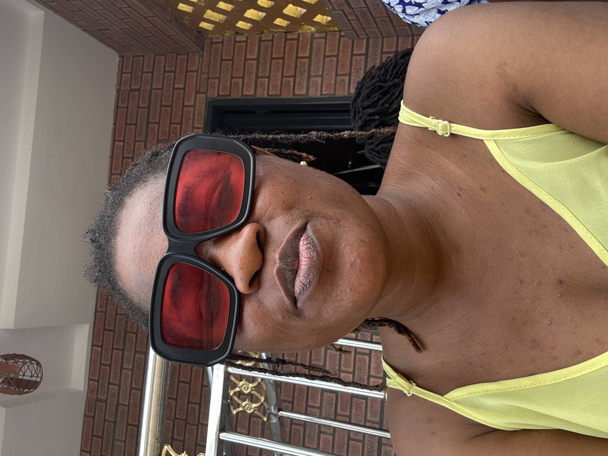 A medium shot shows a Black woman from the chest up, wearing large, square, black-framed sunglasses with red lenses and a light yellow spaghetti-strap top. Her locs are visible on either side of her face, with one strand falling in front of her right eye. Her mouth is slightly open. The background consists of a brick wall and a metal railing with ornate gold detailing. The scene appears to be outdoors, possibly on a porch or balcony, consistent with a residential area in Ajara, Nigeria.