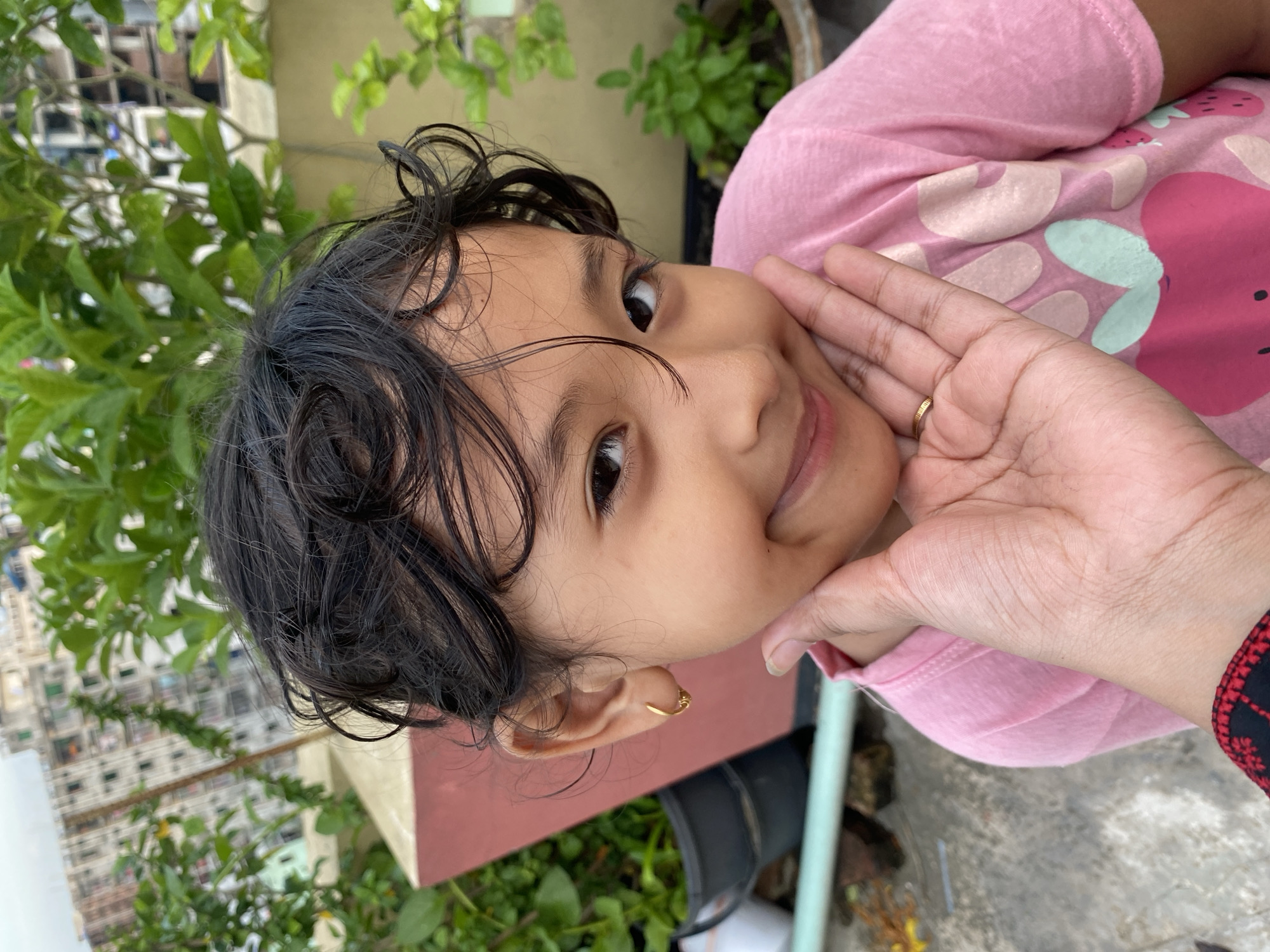 A young girl with dark, curly hair is looking at the camera with a slight smile, her cheek gently held by an adult's hand. She is wearing a pink t-shirt with a strawberry print. The background shows lush green plants and a building, suggesting an outdoor urban setting, possibly a rooftop in Dhaka, Bangladesh. The lighting is soft and natural, indicating it could be daytime.