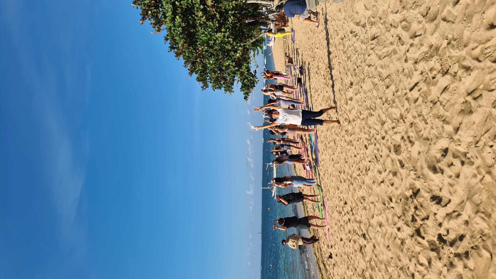 A group of approximately 20 individuals are engaged in a yoga session on a sandy beach. The people are arranged in several rows, facing the ocean. Most are standing and performing yoga poses with their arms raised. Some are wearing athletic attire, while others are in casual clothing. There are yoga mats laid out on the sand. In the background, the ocean stretches out under a clear blue sky. Several boats are visible on the water, along with a red and white lighthouse in the distance. A large tree with green foliage is positioned on the right side of the image, providing shade. The scene appears to be taking place in Denpasar, Indonesia, given the tropical environment and coastal setting. The sand is a light brown color, and the water is a deep blue. The overall atmosphere is peaceful and active, with the participants focused on their exercise.
