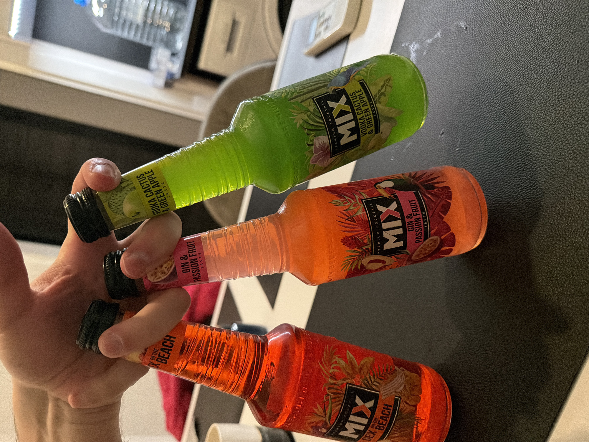 The image shows a hand holding three small bottles of "MIX Premium Cocktail". The bottles are stacked on top of each other, with the bottom bottle labeled "Sex on the Beach", the middle bottle labeled "Gin & Passion Fruit", and the top bottle labeled "Vodka Cactus & Green Apple". The bottles appear to be unopened, with their black caps intact. The background consists of a black textured surface and glimpses of a kitchen interior with cabinets, a water bottle, and other items. There is no information in the image to determine if this image was taken in Plovdiv, Bulgaria.