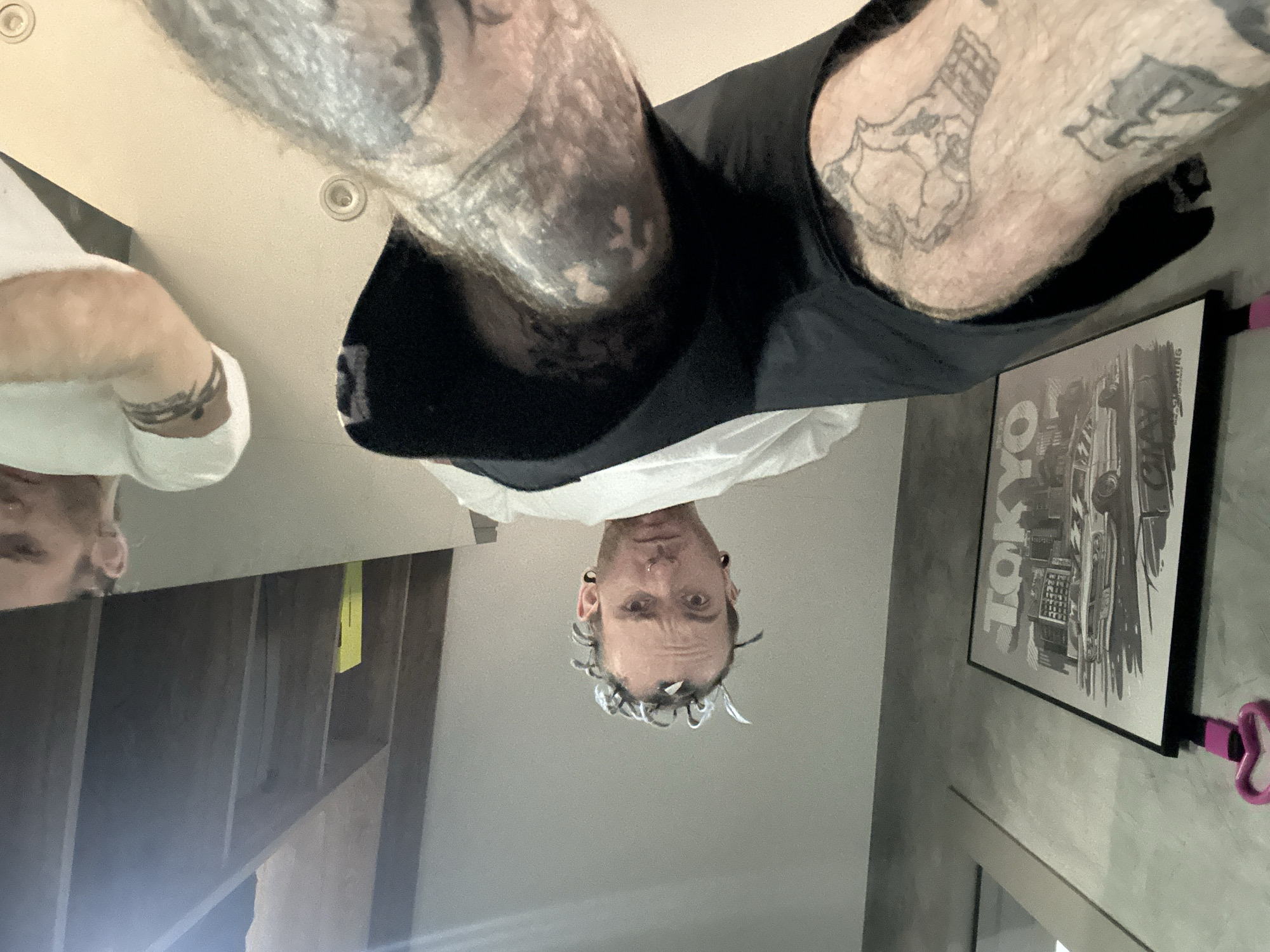 The image presents an upside-down, low-angle view of a person with tattoos on their legs and arms. They are wearing black shorts and a white t-shirt. The person has short hair with what appears to be small metal objects attached to it. Their face is visible, showing a neutral expression.
In the background, there is a framed black and white poster with the word "TOKYO" visible. Other elements in the background include a wooden structure, a light switch, and a pink object.
There is no evidence to suggest that this photo was taken in Khimki, Russia.