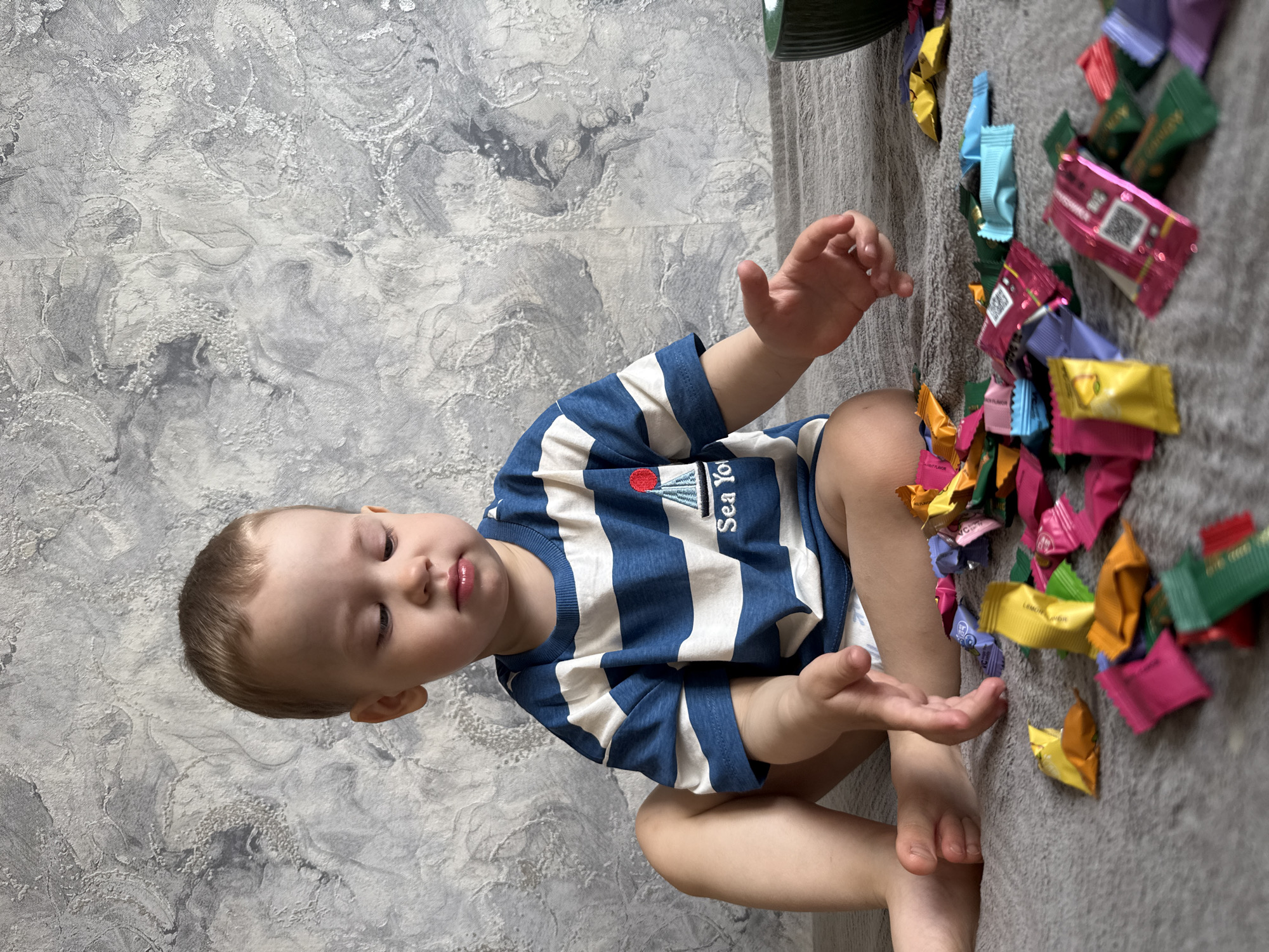 A young child, likely a toddler, is sitting on a grey, textured surface, possibly a blanket or carpet, in an indoor setting. The child has light brown hair, fair skin, and dark eyes, looking down and slightly to their right. They are wearing a blue and white striped short-sleeved t-shirt with a small red sailboat emblem and partially visible text "Sea Yo..." (likely "Sea Voyage" or "Sea Story") on the left chest.
The child's hands are open and slightly raised, suggesting interaction or anticipation with the numerous small, brightly colored candy wrappers scattered on the grey surface to their right. The wrappers are in shades of pink, yellow, green, blue, purple, and orange. Visible text on some yellow wrappers reads "LEMON FLAVOR." A pink wrapper shows a square code resembling a QR code and some small, indistinct text. A dark green wrapper also features some small, indistinct text, possibly in an East Asian script.
Behind the child is a light grey wall with a textured, abstract pattern resembling marble or a damask design. The overall scene conveys a moment of playful interaction with sweets. The child's expression is calm and focused. The image is taken indoors, with no discernible weather or specific time of day cues other than sufficient lighting. This scene is situated in Rusanovo, Russia.