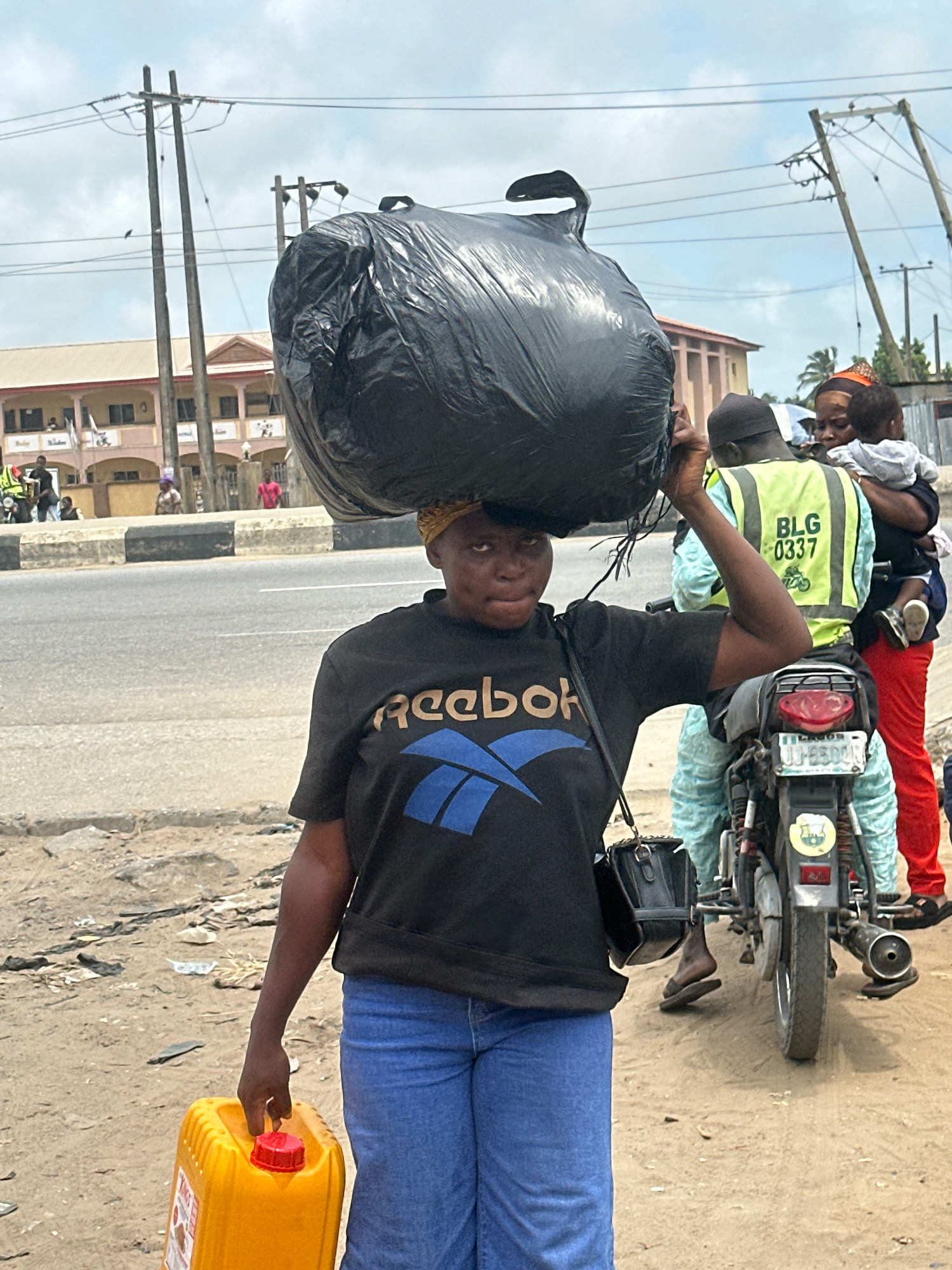In the city of Ajara, Nigeria, a woman walks carrying a large black plastic bag balanced on her head. She is wearing a black t-shirt with a blue Reebok logo and blue jeans. In her right hand, she holds a bright yellow plastic jerrycan with a red cap. Behind her, a person wearing a reflective yellow vest with "BLG 0337" printed on it rides on the back of a motorcycle. This person appears to be holding a baby. Another person in red pants stands near the motorcycle. The scene takes place on a dirt shoulder next to a paved road. In the background, there are buildings and utility poles. The sky is overcast.