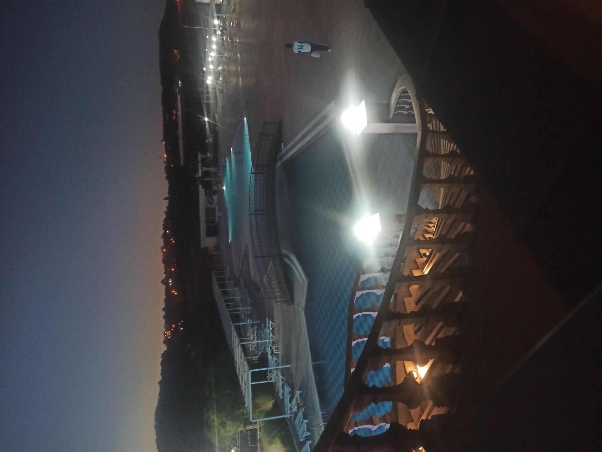 An aerial, full shot captures a resort-style swimming pool area at dusk. The scene is illuminated by artificial lighting from the pool itself, overhead lamps, and distant city lights. In the foreground, a dark, ornate wooden balcony railing curves into the frame, leading down a dimly lit staircase.
The main focus is the spacious, multi-tiered swimming pool, its turquoise water reflecting the lights. A arched pedestrian bridge spans across one section of the pool. Surrounding the pool are rows of white lounge chairs under white canopies, suggesting a place for relaxation. Further in the distance, more pools and seating areas are visible, along with a building structure.
A lone individual, a male wearing a white t-shirt with the number '14' on the back and dark shorts, walks away from the viewer across the paved area near the pool. The background depicts a dark, hilly landscape dotted with glowing lights from a settlement. The sky is a gradient of deep blue and faint orange, indicating twilight.