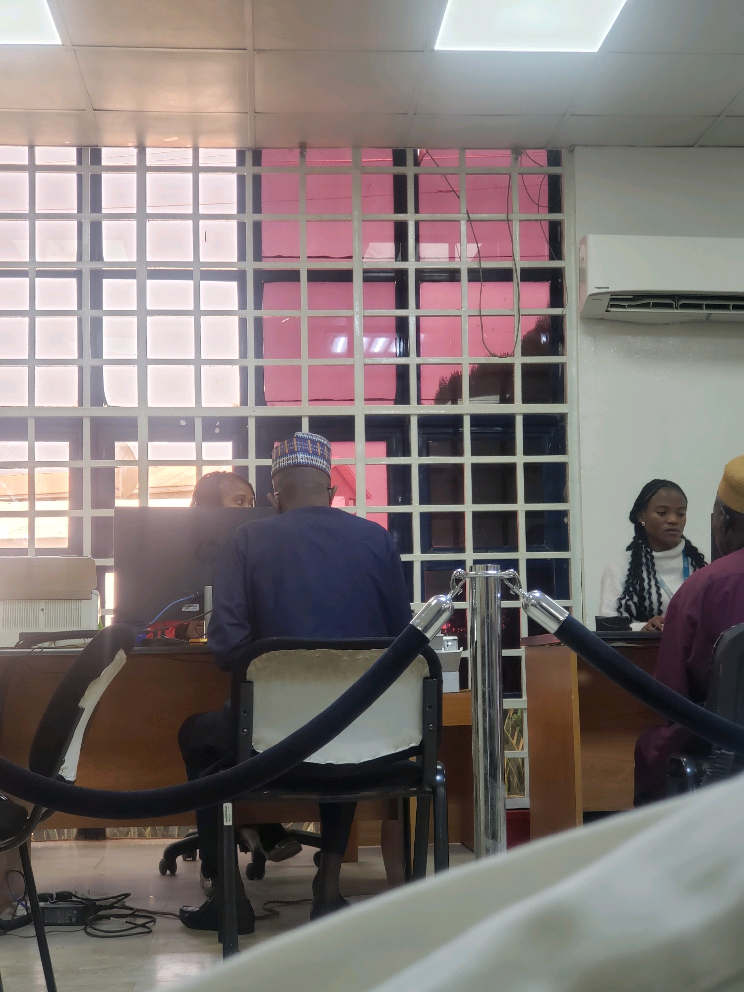 The image depicts an indoor scene, possibly a government office or a bank, in Kakuri Gwari, Nigeria. In the foreground, a black velvet rope barrier is visible, suggesting a queue or waiting area. Behind the barrier, a person wearing a dark blue traditional outfit and a patterned cap is seated at a desk, facing away from the camera. They are looking towards a computer monitor. To their left, another individual, partially obscured by the monitor, appears to be interacting with them.
Further back, another desk is occupied by a woman with braided hair, wearing a white turtleneck sweater. She is also looking towards something out of frame. To the far right, the shoulder and head of another person, wearing a burgundy garment and a light-colored cap, are visible.
The environment features a white ceiling with grid lighting and a window with a grid pattern, behind which a pinkish hue is visible, suggesting tinted glass or a colored wall. An air conditioning unit is mounted on the wall. The flooring appears to be tiled. The overall impression is one of an organized, albeit somewhat dated, administrative setting.